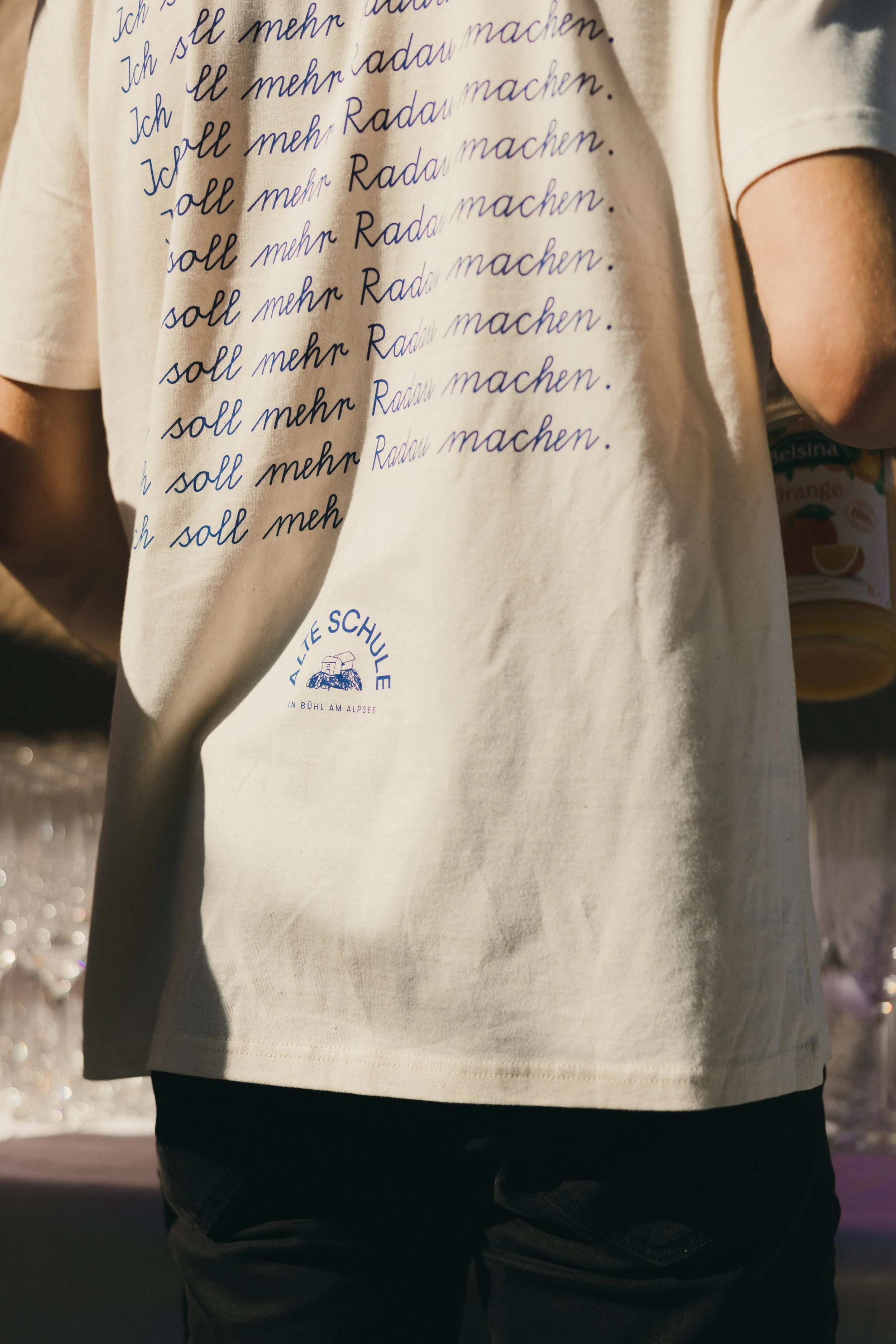 Person trägt weißen T-Shirt mit blauer Handschrift, die wiederholt 'Ich soll mehr Rad fahren' sagt, und schwarzen Hosen, im Hintergrund Wasserstrahlen.