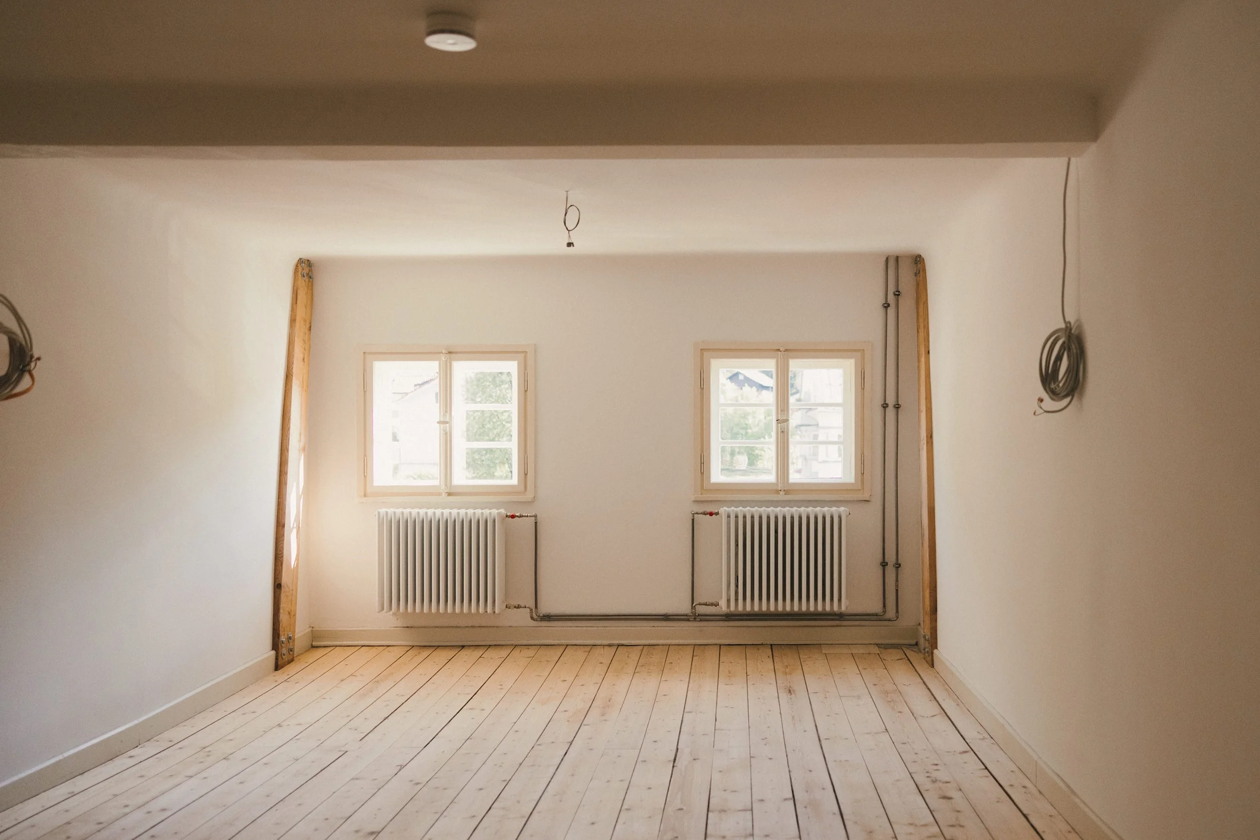 Ein unmöbliertes Zimmer mit weißen Wänden, Holzfußboden und zwei Fenster, darunter zwei Heizkörper. Es sind Kabel an den Wänden und eine Deckenleuchte.