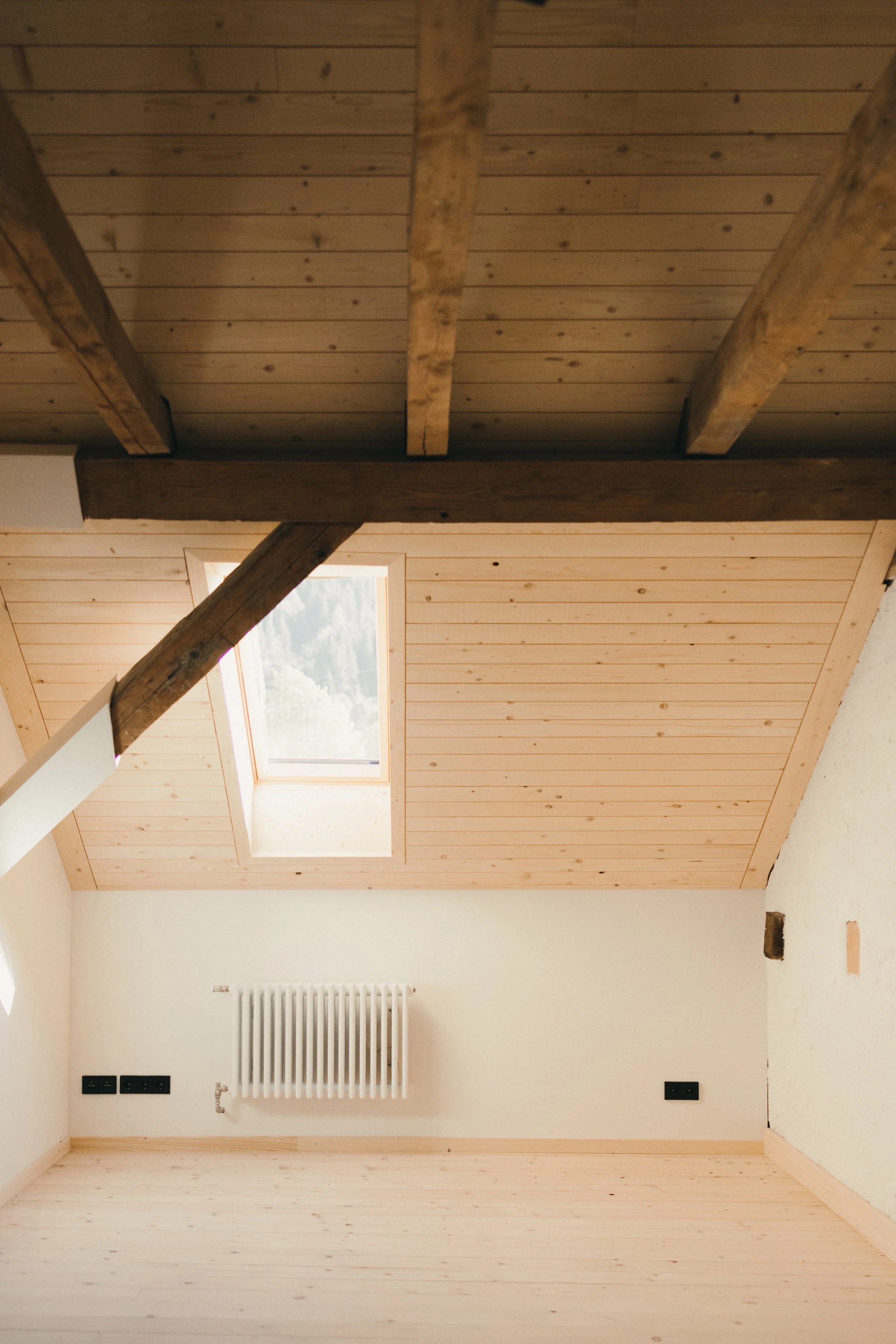 Innenaufnahme eines Raumes mit Holzbalkendecke und einem Dachfenster, weiße Wände und Boden, Heizkörper und elektrische Anschlüsse sichtbar.
