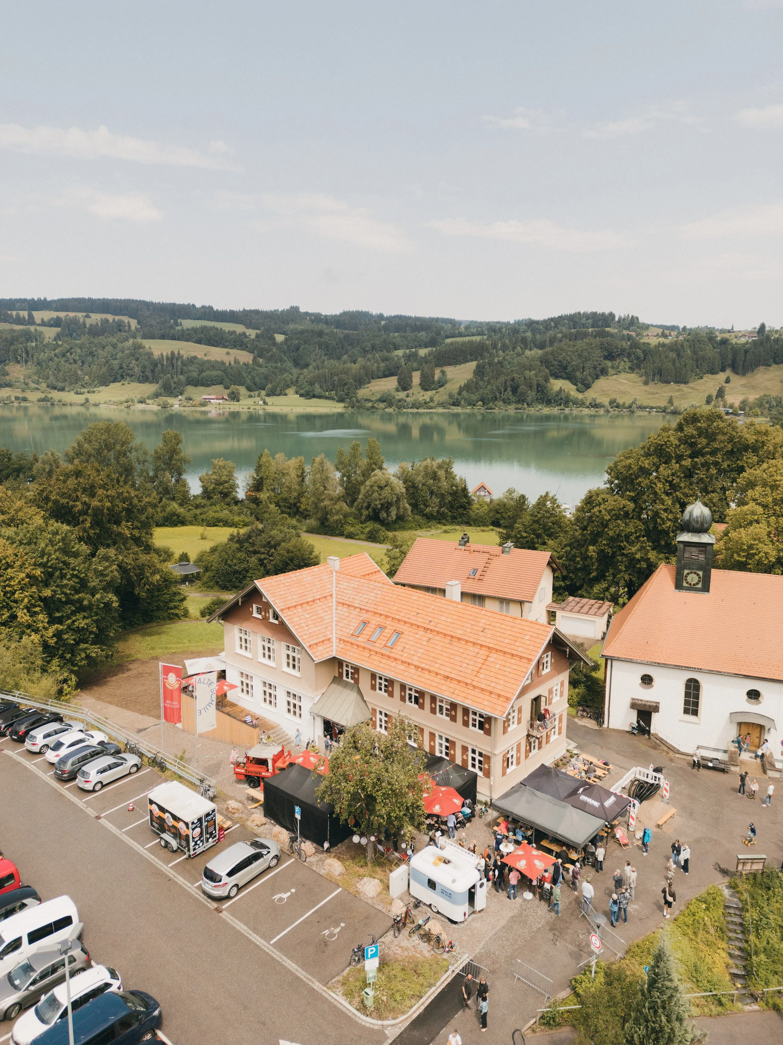 Blick auf ein Fest mit mehreren Zelten und Ständen vor einem historischen Gebäude mit rotem Dach und Uhrturm, Menschen und Fahrzeuge im Vordergrund, umgeben von Bäumen und Hügeln.