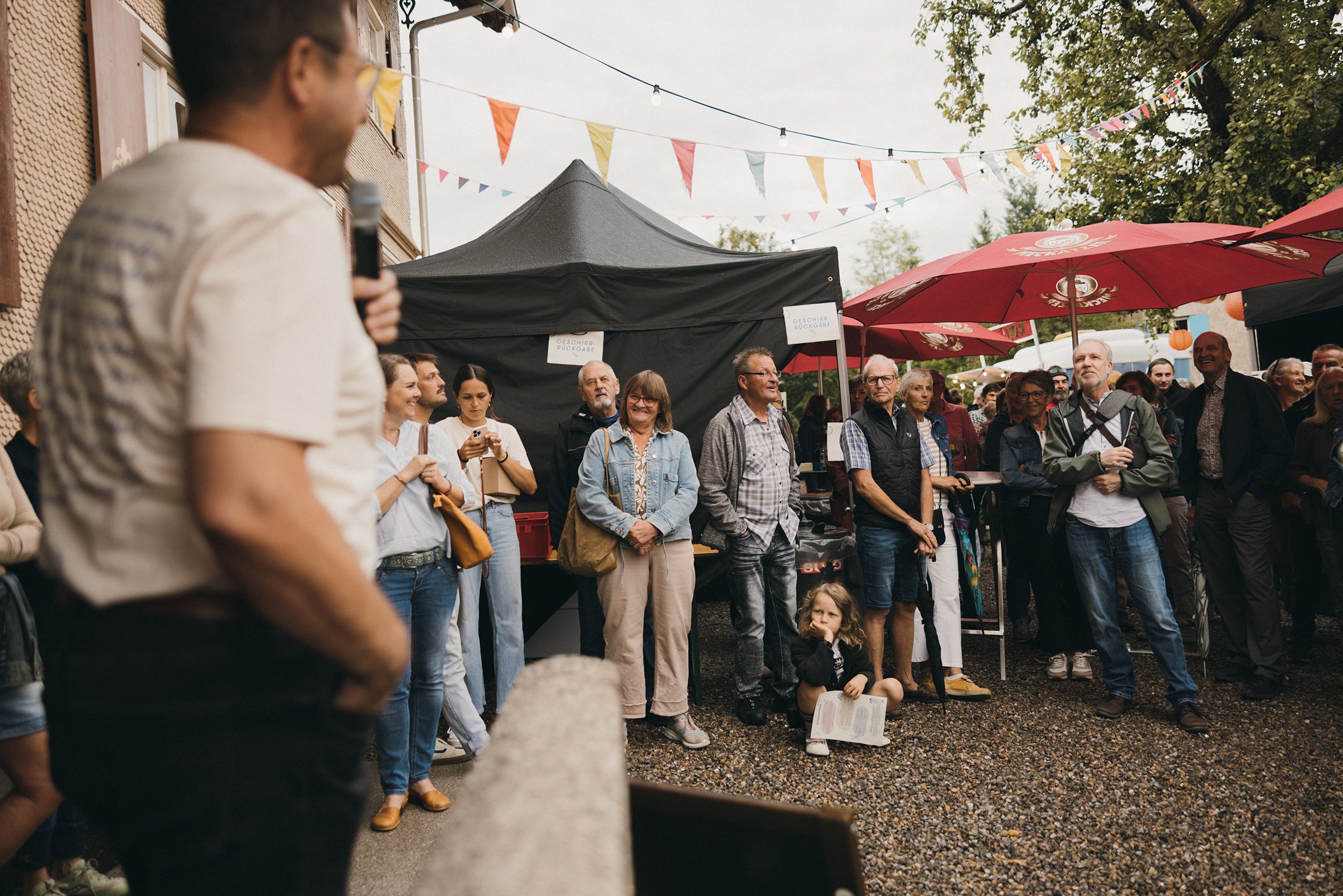 Menschen versammeln sich bei einem Straßenfest unter bunt geschmückten Flaggen. Es scheint eine Unterhaltung oder eine Präsentation zu geben, während die Leute im Freien unter einem großen roten Sonnenschirm stehen.