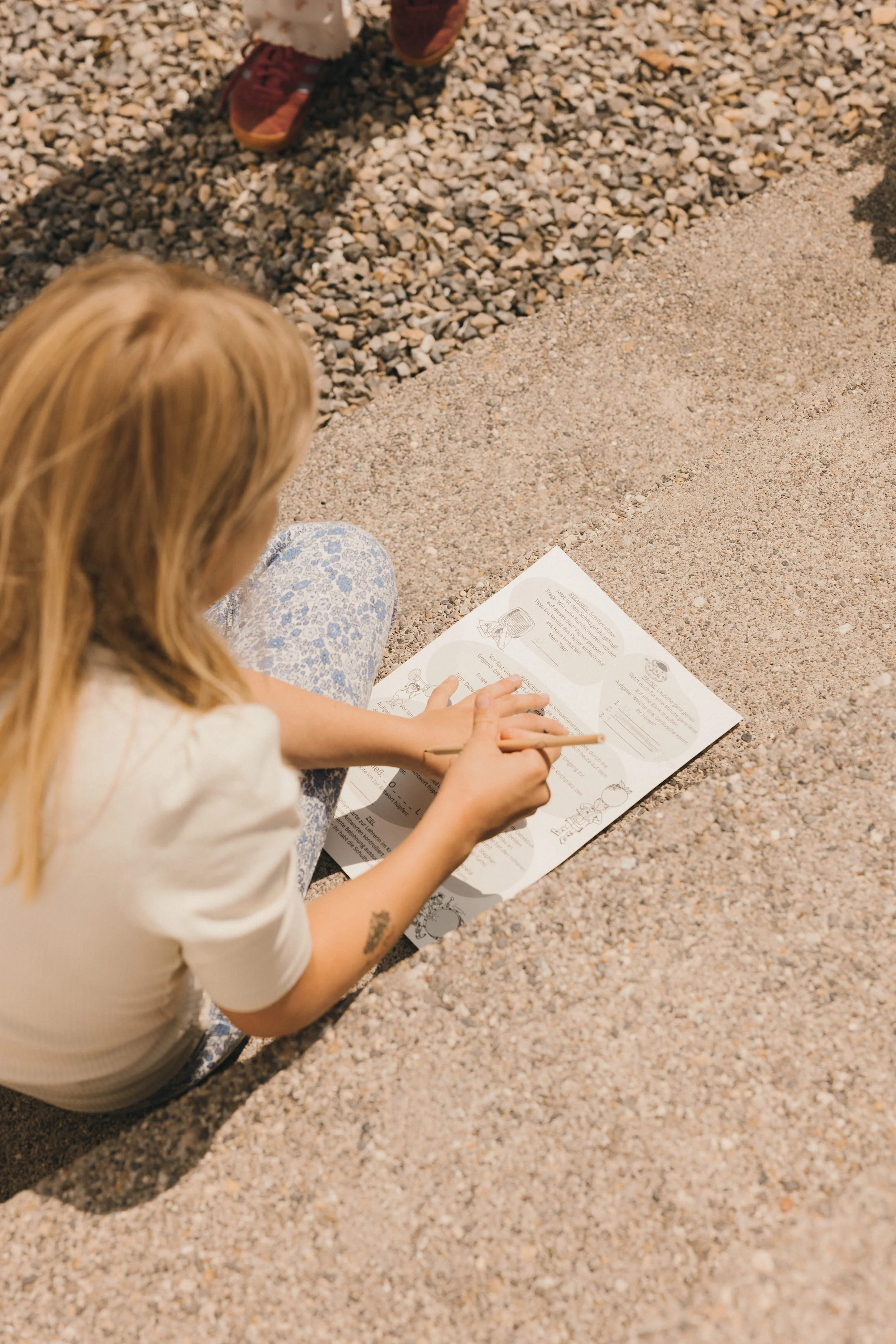 Mädchen liest ein Buch im Freien auf einer Beton- und Kiesfläche.