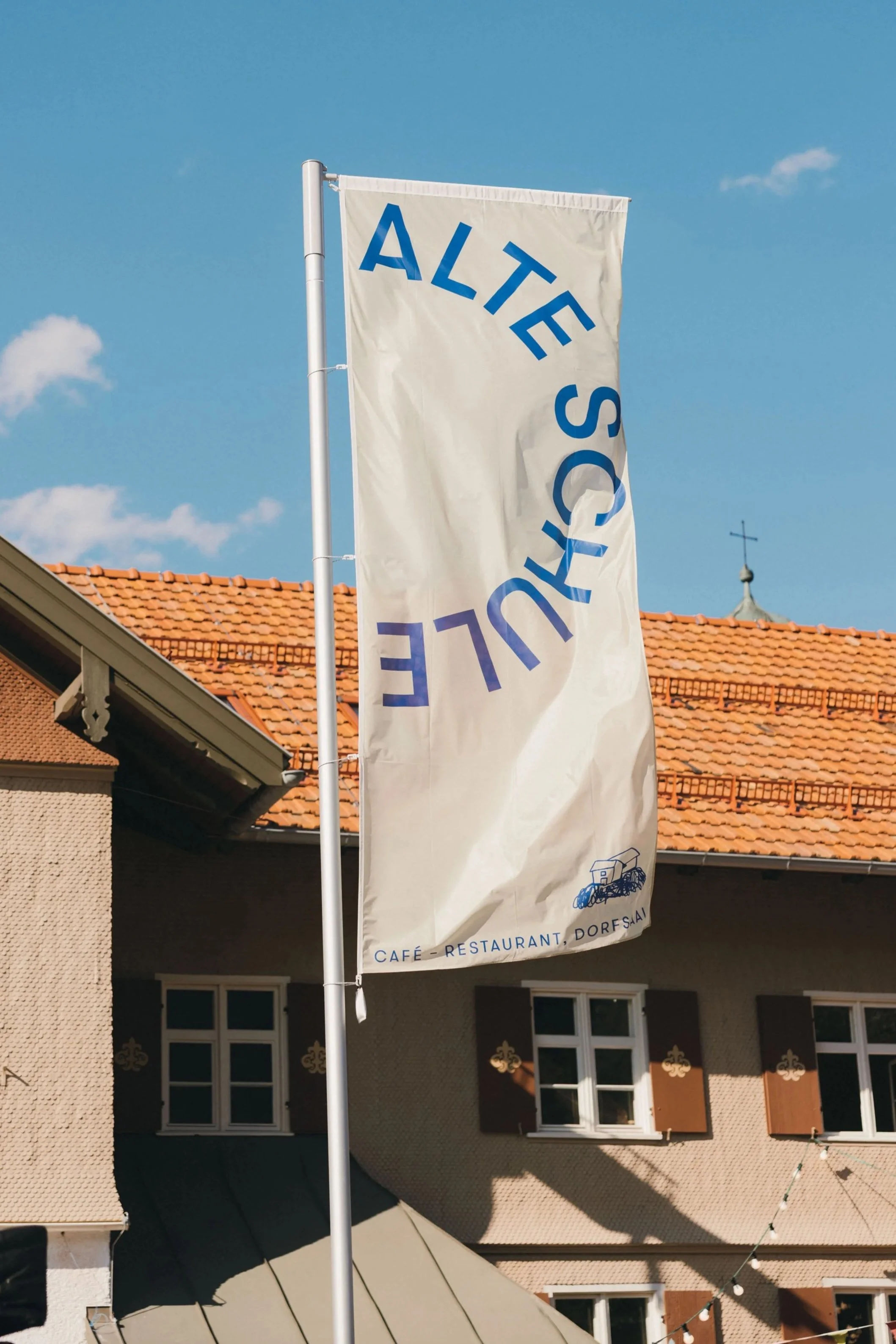 Ein weißes Banner mit blauer Schrift mit der Aufschrift 'ALTE SO.' Es ist an einem weißen Fahnenmast befestigt und im Hintergrund sind Dächer mit roten Ziegeln, Fenster und eine kleine Turmspitze mit Kreuz zu sehen, bei sonnigem Himmel.