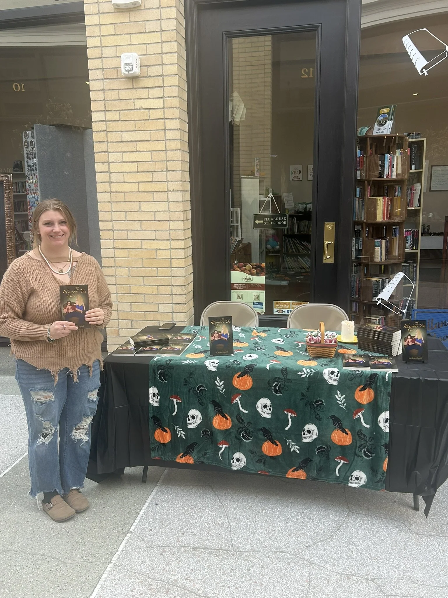 A woman standing next to a table covered with a Halloween-themed tablecloth featuring skulls, pumpkins, mushrooms, and black cats. The table has copies of a book titled "It Was Me,