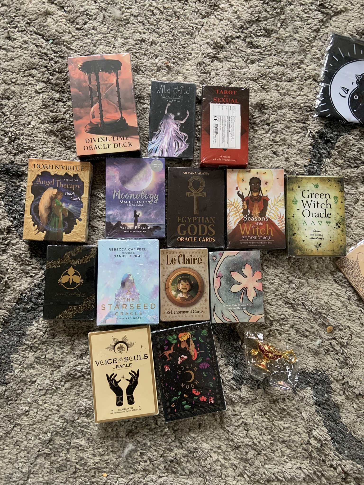 A collection of tarot and oracle card decks arranged on a gray carpet, including decks titled 'Divine Time Oracle Deck,' 'Wild Child Oracle Cards,' 'Tarot of Sexual,' 'Angel Therapy Oracle Cards,' 'Moonology Manifestation Oracle Cards,' 'Egyptian Gods Oracle Cards,' 'Seasons of the Witch,' 'Green Witch Oracle,' 'The Starseed Oracle,' '36 Lenormand Cards,' 'Voice of the Souls Oracle,' and others. There is also a gold-colored charm and a clear plastic bag with a decorative item.