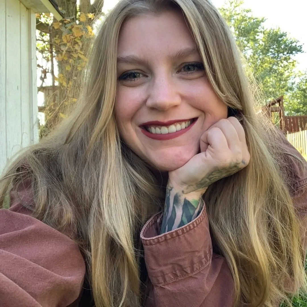 A young woman with long blonde hair, smiling, resting her chin on her hand, outdoors with trees and a wooden fence in the background.