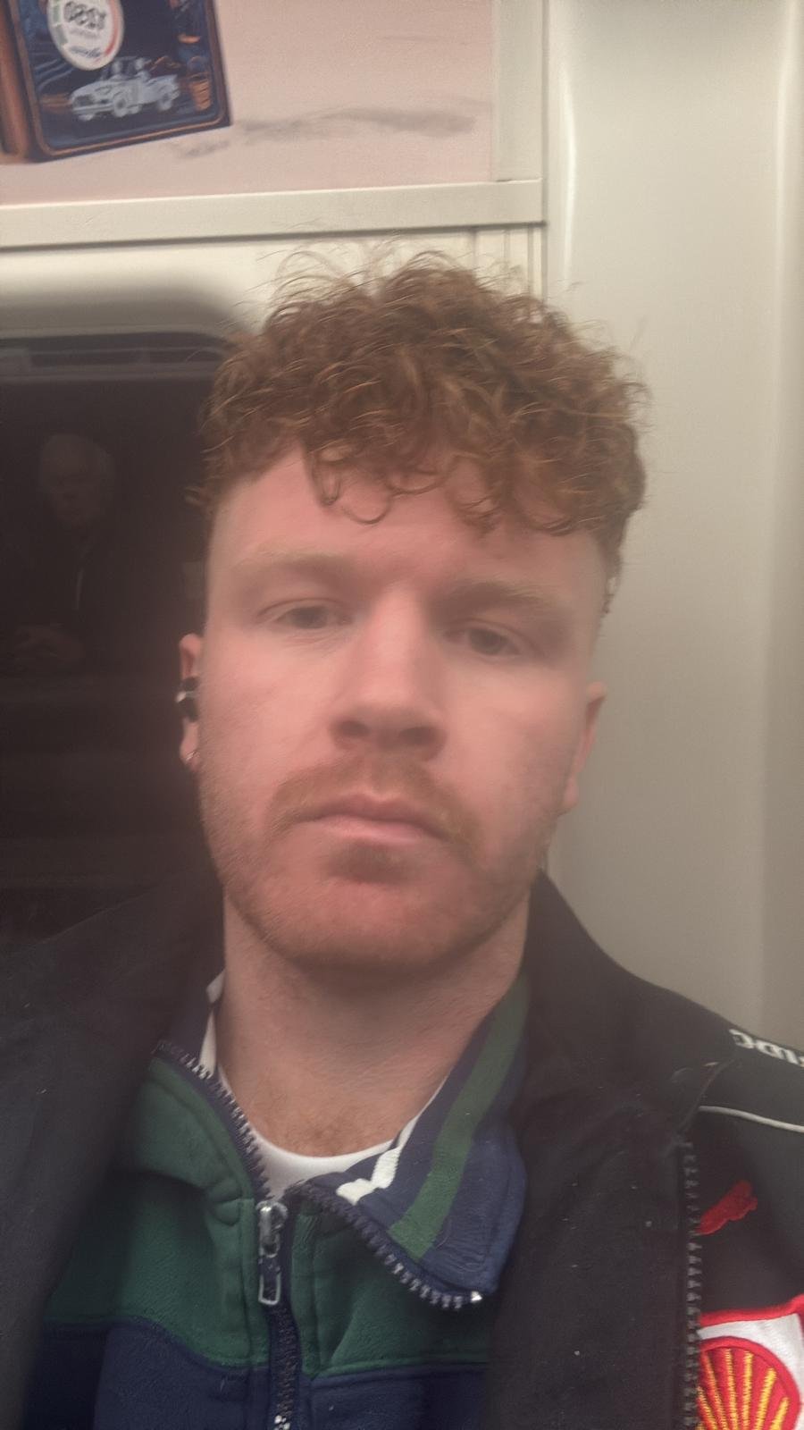 Young man with curly red hair taking a selfie, with a serious expression, wearing a green and blue jacket with a zipper, sitting in a vehicle or train.
