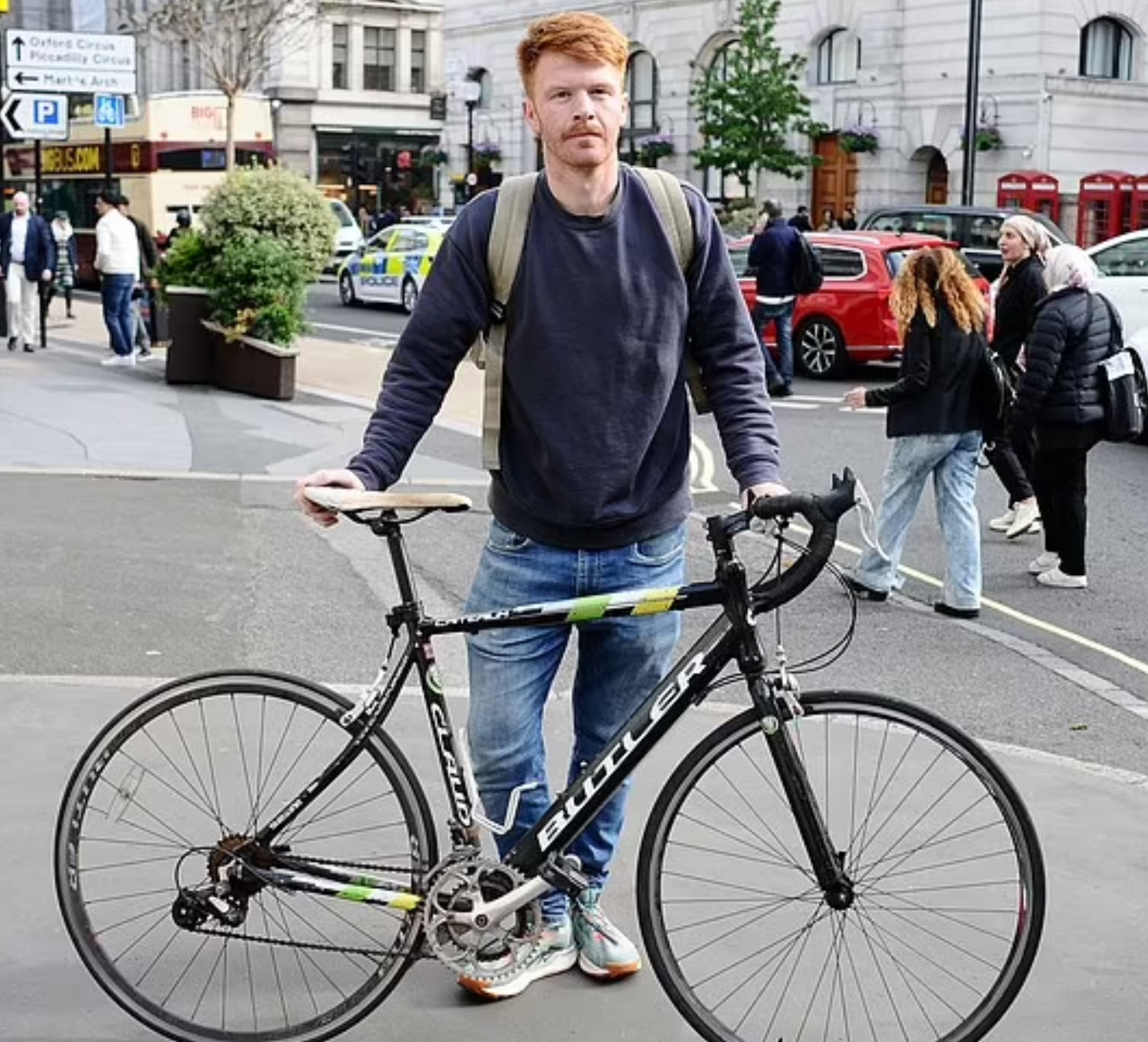 A man with red hair and a beard standing with a black and white Trek bicycle on a city street. He wears a dark sweatshirt, blue jeans, sneakers, and carries a backpack. Pedestrians walk by and cars are visible in the background.