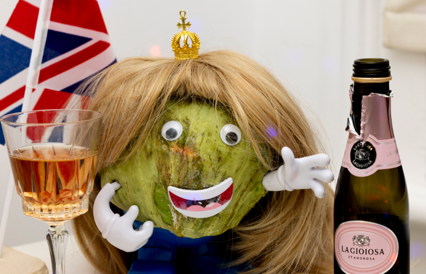 A green coconut with a face, wearing a gold crown, with a blonde wig, googly eyes, a mouth with a big smile, white gloves, and blue clothing, sitting among a glass of rosé wine, a bottle of La Gioiosa sparkling wine, and a British flag in the background.