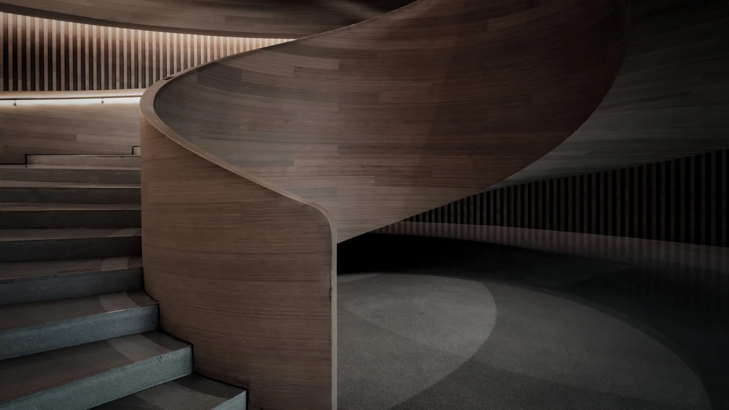 Interior staircase with wooden handrail and dark steps, curved wooden wall, and striped wall panels in a modern architectural space.