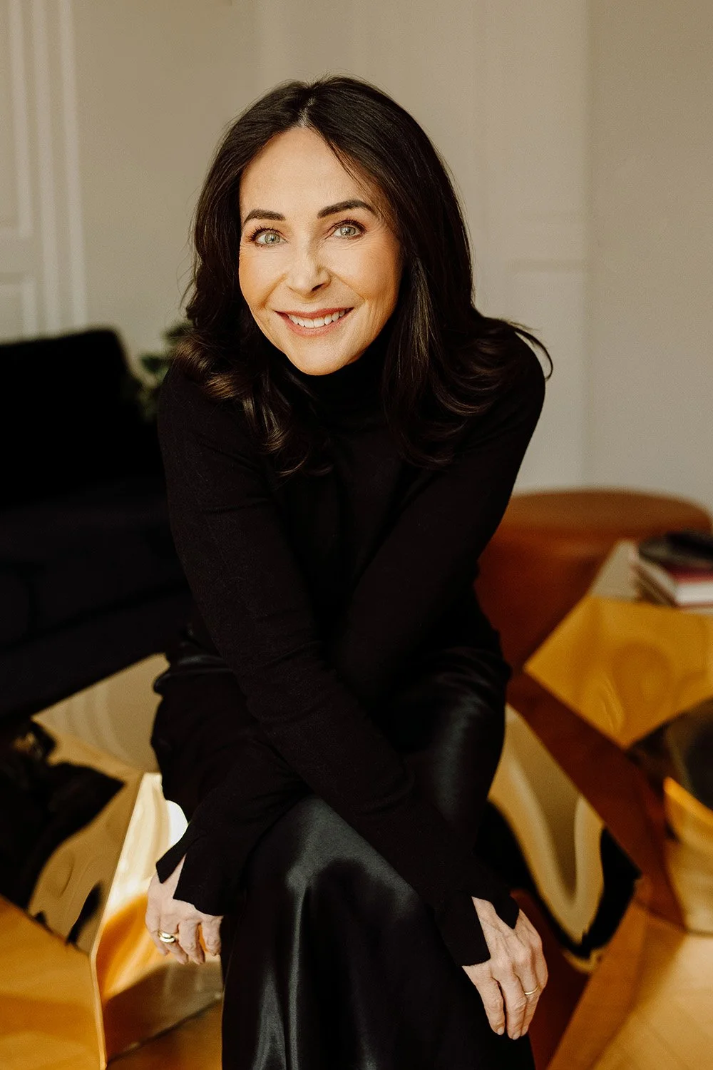 A woman with dark hair and blue eyes, smiling, wearing a black turtleneck and leather pants, sitting on a chair in a cozy indoor setting.