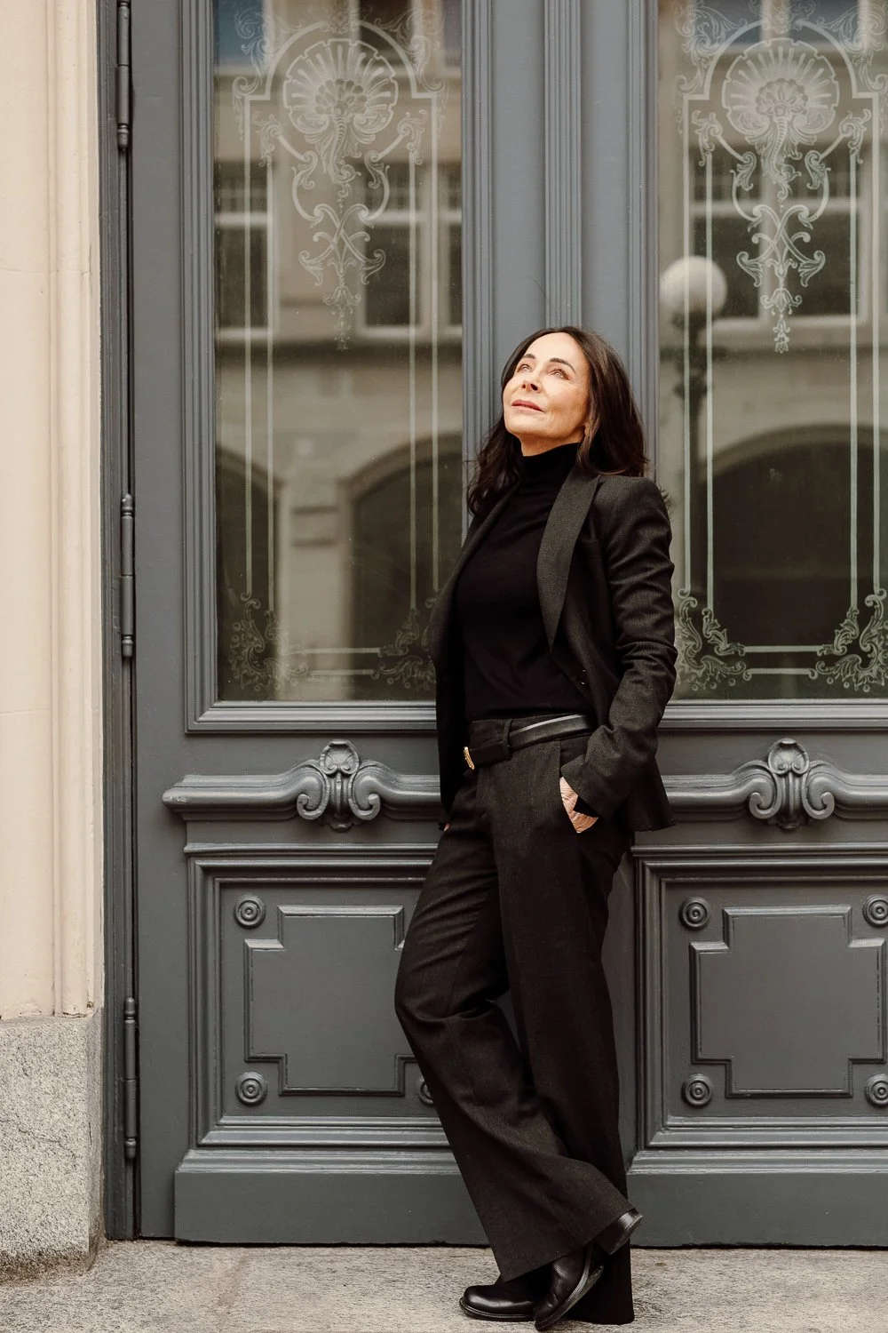 A woman in a black turtleneck and black blazer poses against an ornate gray door, looking upward with her hands in her pockets.