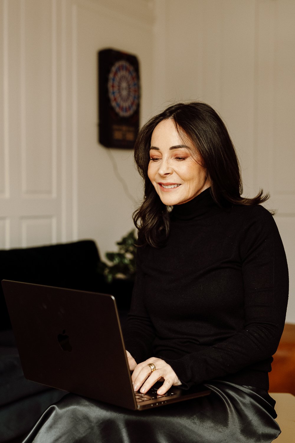 A woman with shoulder-length dark hair and a black turtleneck shirt sitting at a table, smiling while working on a laptop in a cozy room with a dartboard on the wall in the background.