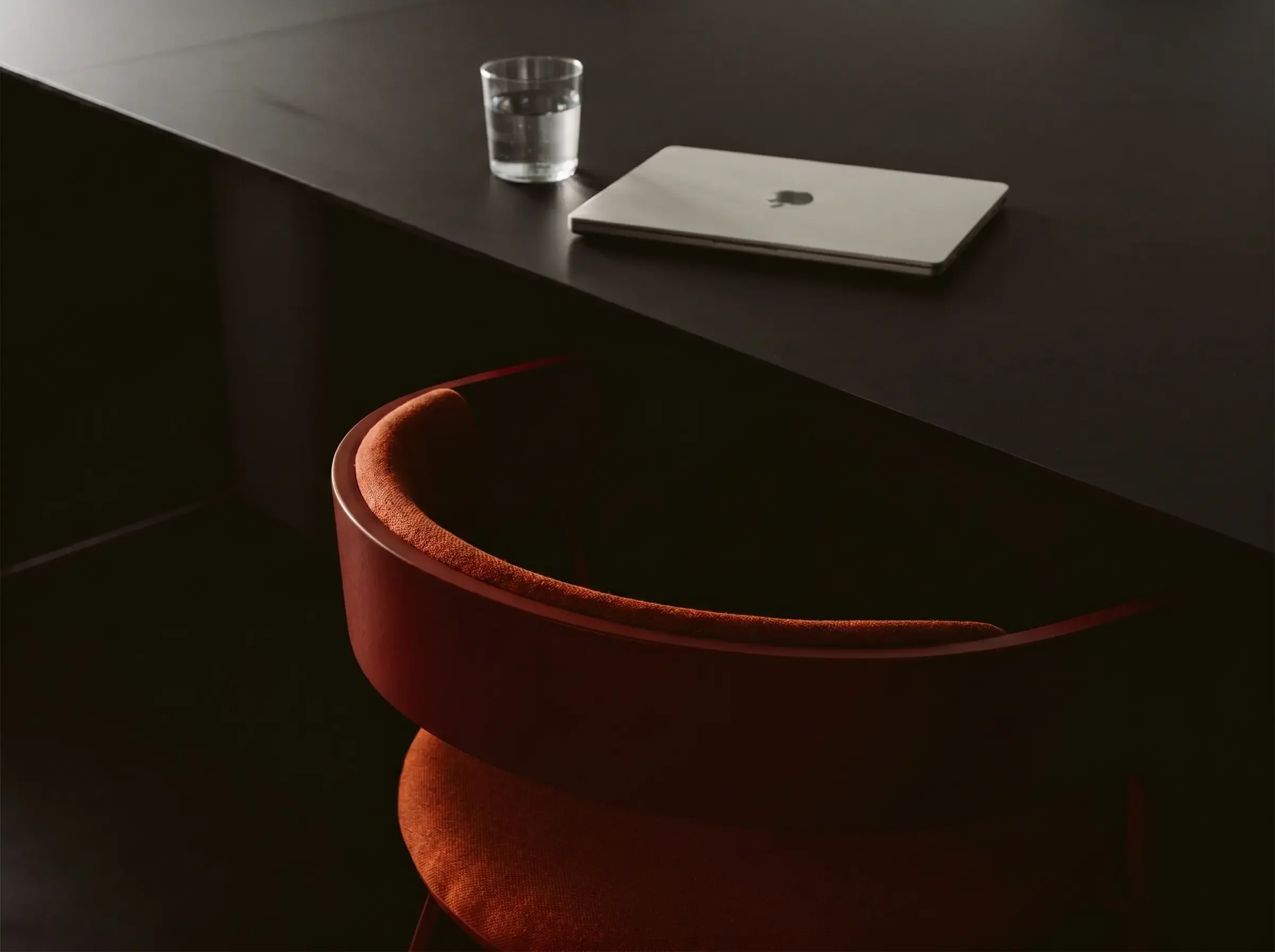 A black table with a glass of water, a closed silver laptop, and a red velvet chair underneath, in a dimly lit setting.