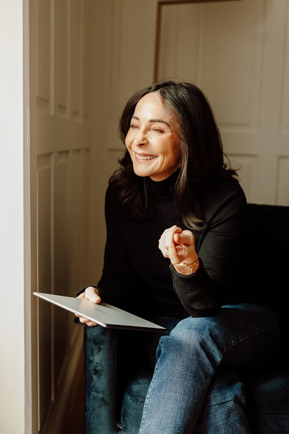 A woman with shoulder-length dark hair, smiling and laughing, sitting on a dark upholstered chair indoors. She is wearing a black turtleneck and blue jeans, holding a tablet in her left hand, and has a gold watch on her left wrist.
