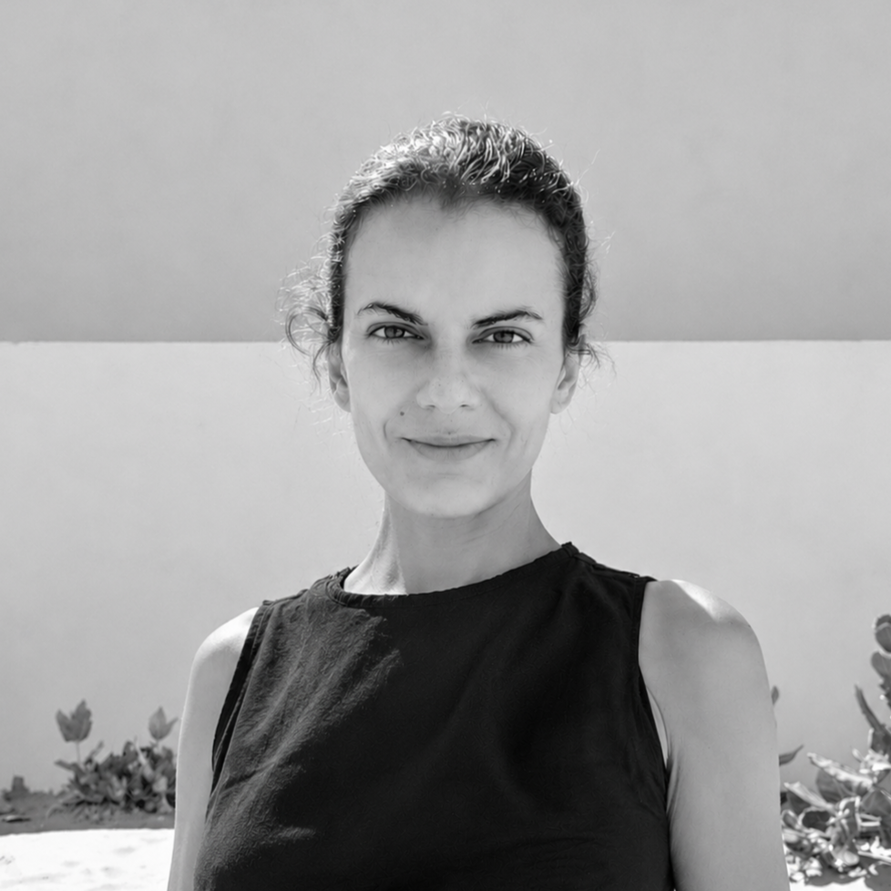 Black-and-white photo of a woman with dark hair pulled back, wearing a sleeveless black shirt, standing outdoors against a plain wall.
