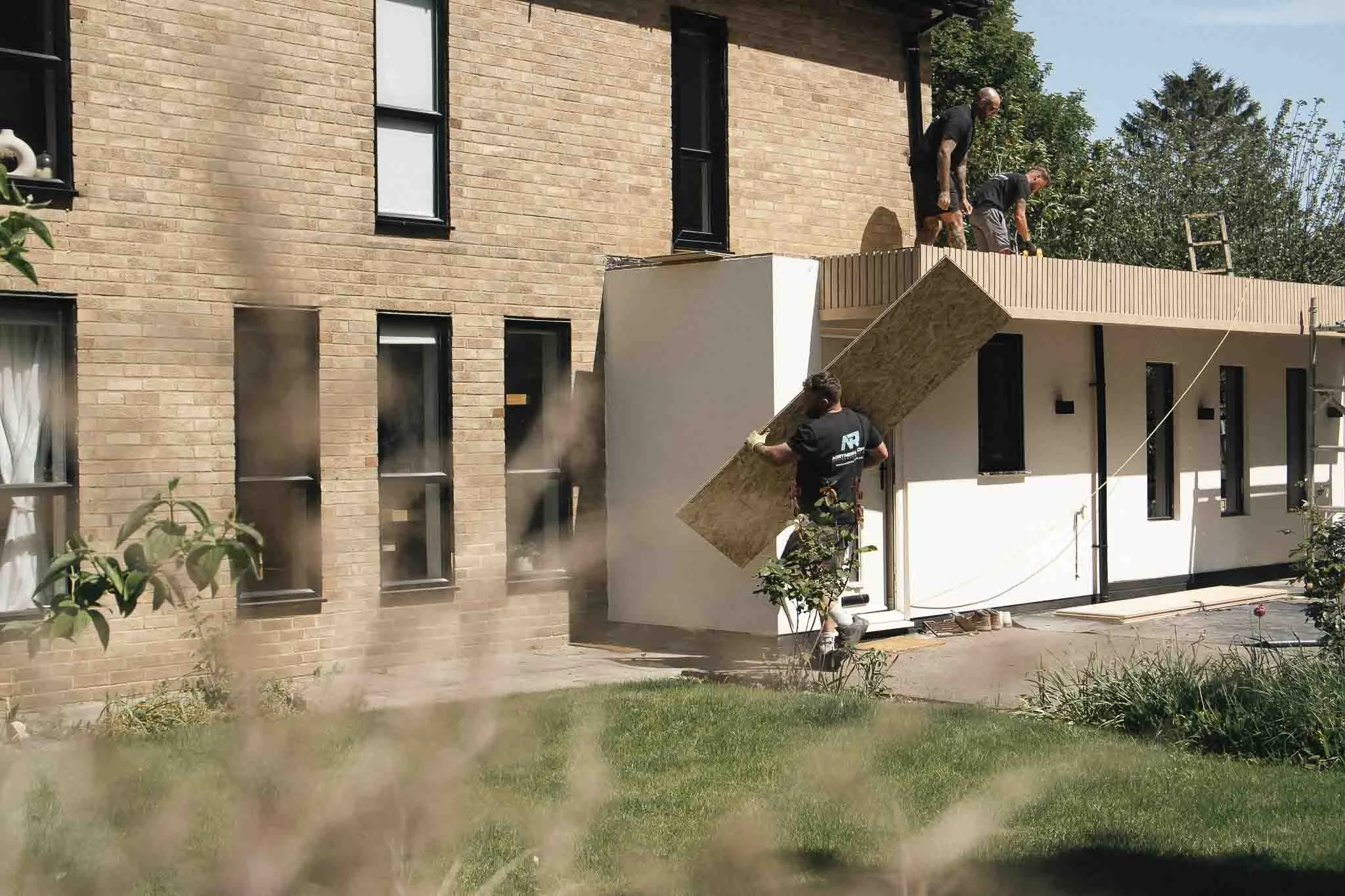 Workers installing a balcony or deck on a brick building. One worker is on the balcony railing, another is on the ground passing materials, and a third is near the structure. The building has multiple windows, and there are trees and a ladder in the background.