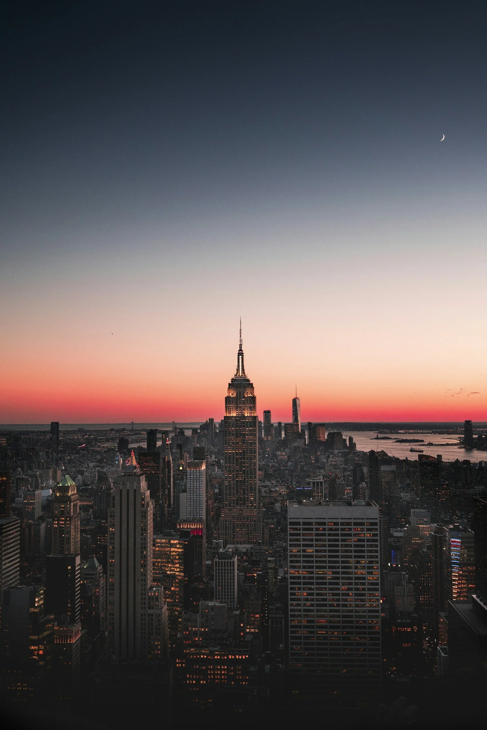 Nighttime cityscape featuring the Empire State Building in New York City with a colorful sunset sky and a crescent moon.