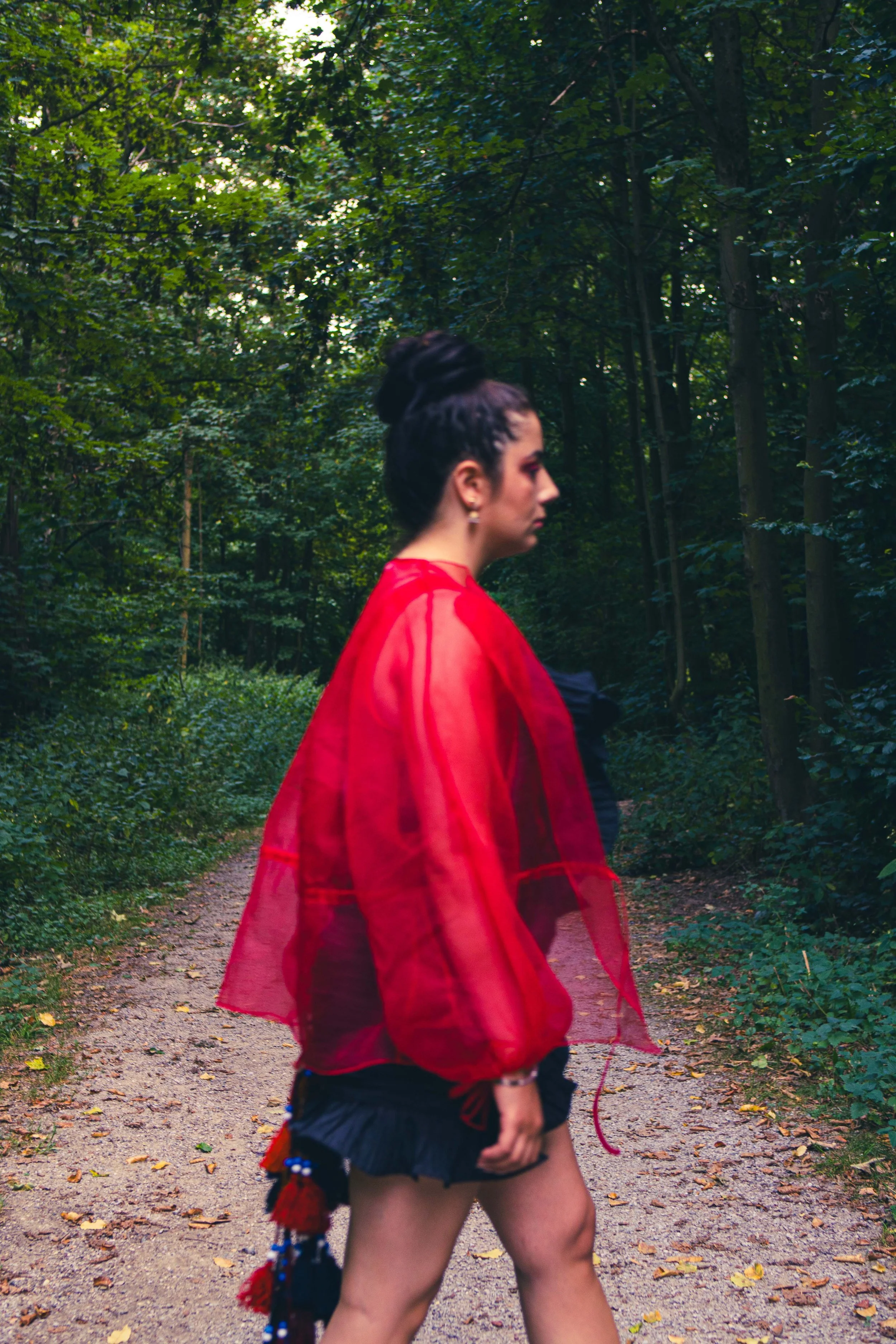 Frau in einem roten transparenten Kleid geht auf einem Waldweg, umgeben von Bäumen und Blättern auf dem Boden.