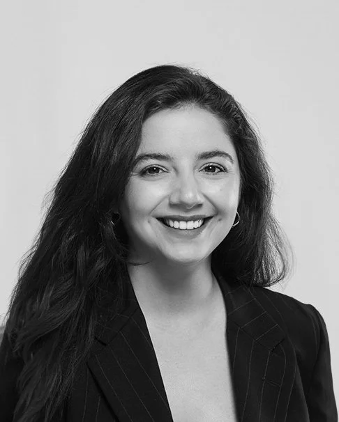 Schwarz-weißes Portrait einer lächelnden jungen Frau mit langen, dunklen Haaren, in Businesskleidung vor hellem Hintergrund.