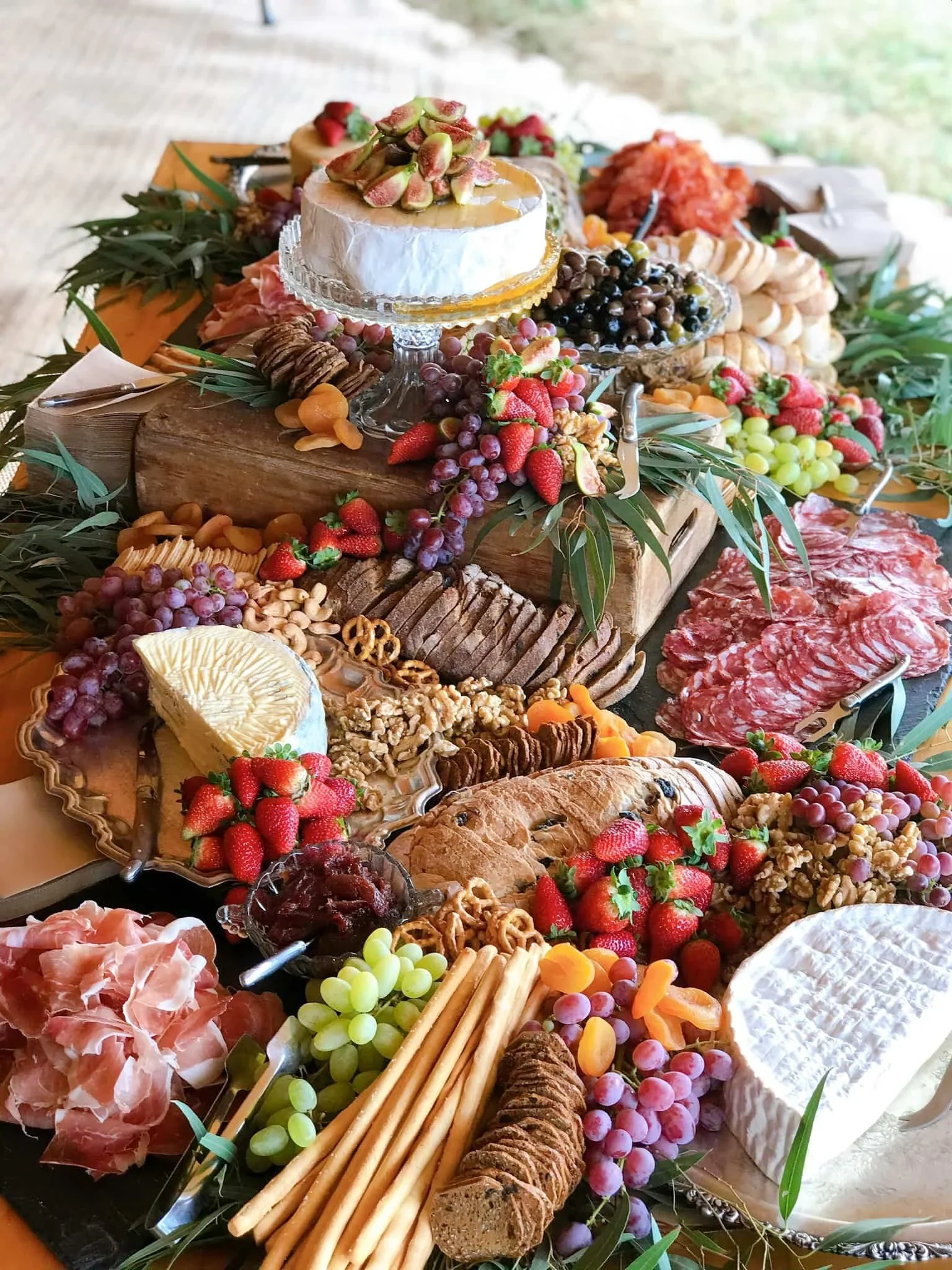 A spread of various cheeses, grapes, meats, crackers, and fresh fruit arranged on a table. The centerpiece features a round cheese with pistachios on top, surrounded by charcuterie, bread, and an assortment of fruits and nuts.