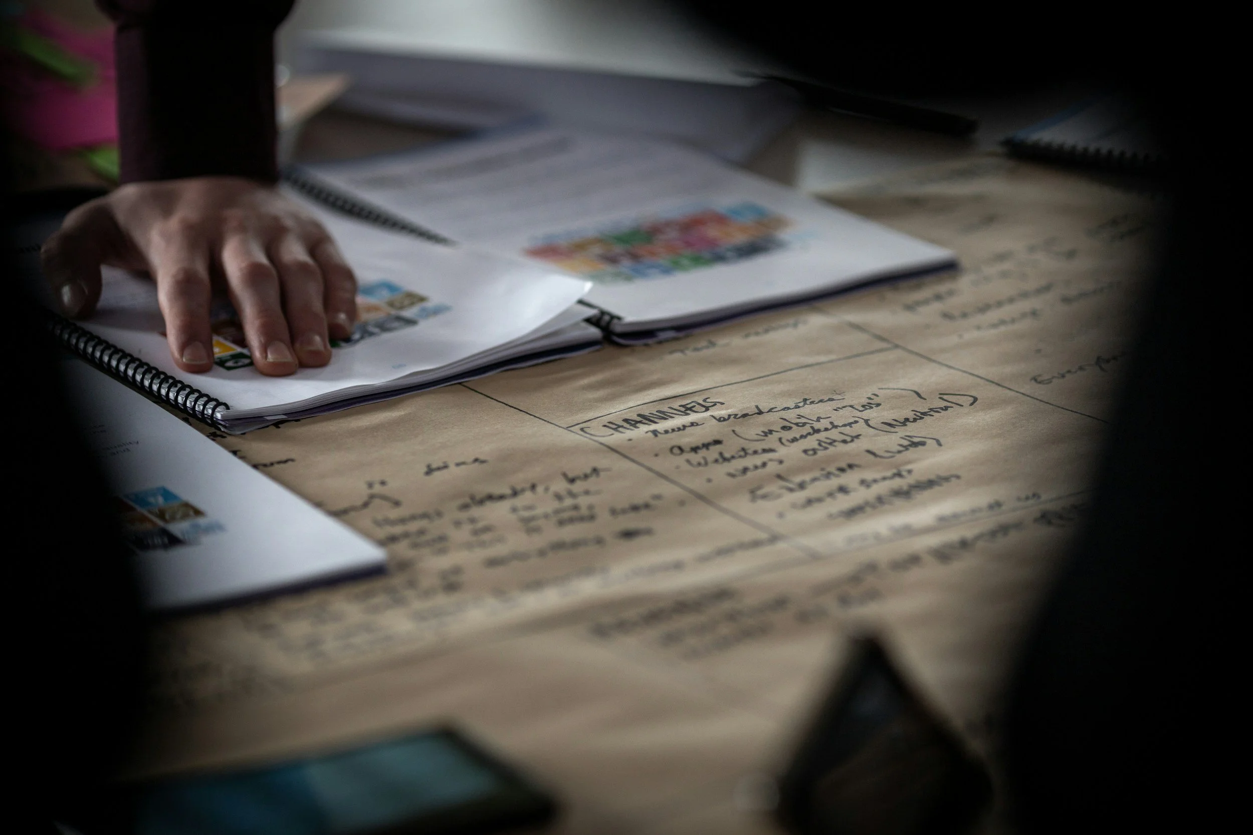 A hand resting on an open spiral-bound notebook on a cluttered desk, with handwritten notes and colorful stickers or images. The scene is dimly lit with shadows around the edges.