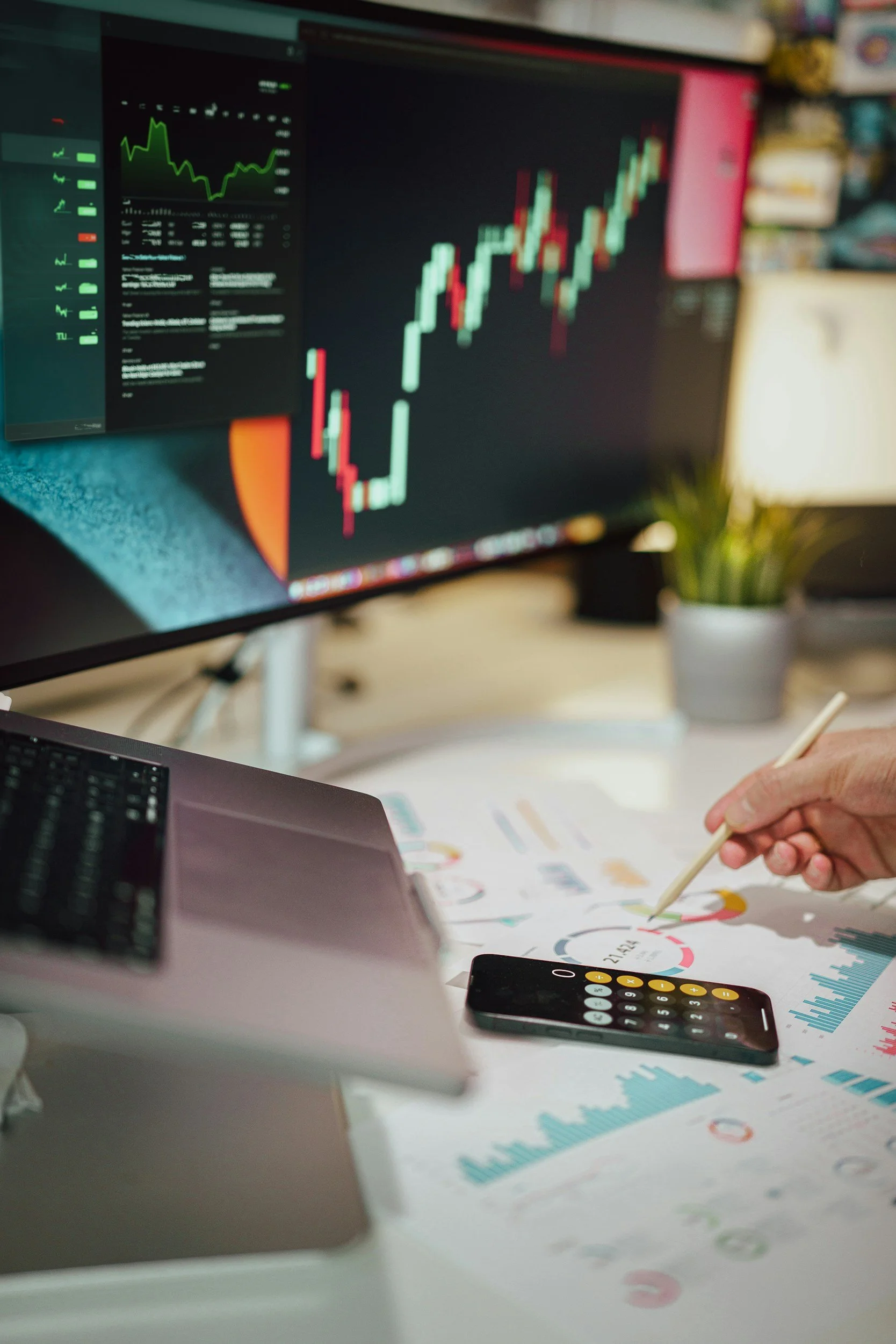 A computer monitor displaying stock market charts and graphs with red and green candlesticks, a hand holding a stylus pointing at printed data and charts on a desk, a calculator, a laptop, and a smartphone.