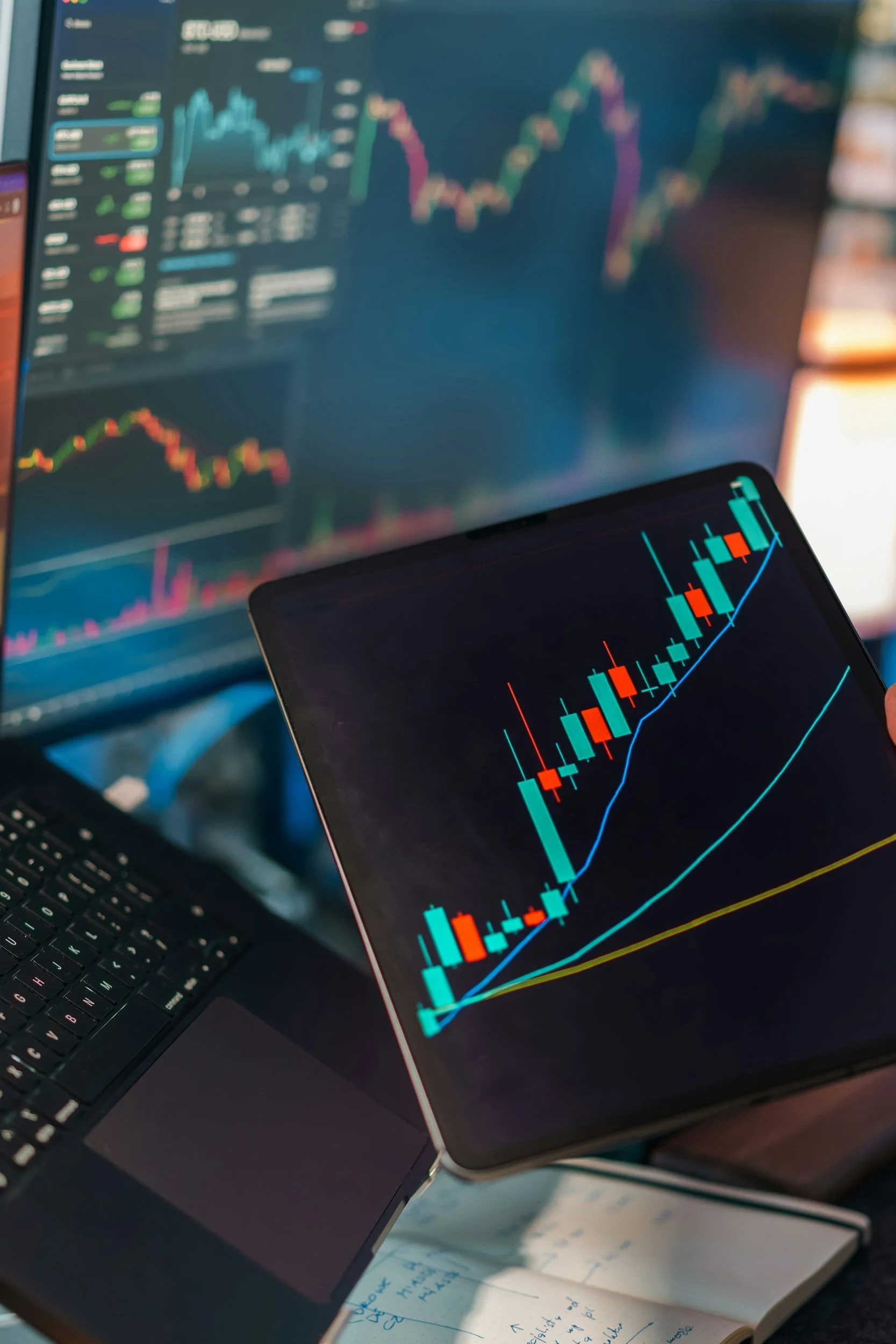 Close-up of a computer monitor and a smartphone displaying stock market charts with candlestick patterns, with a keyboard and handwritten notes on paper in the background.