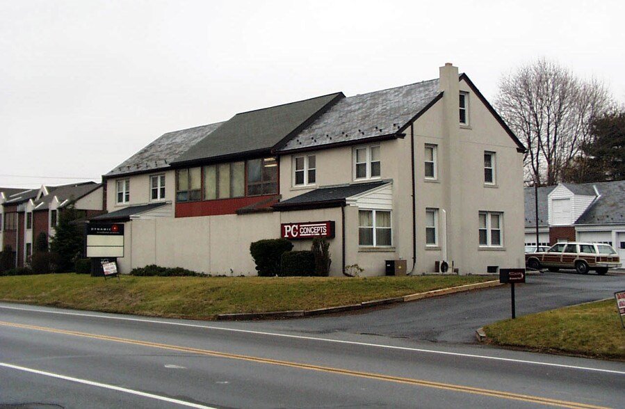 A two-story commercial building with a white exterior, featuring a sign that says 'PC Concepts,' and nearby parking lots with cars. The building has multiple windows and is situated along a street with traffic lanes.