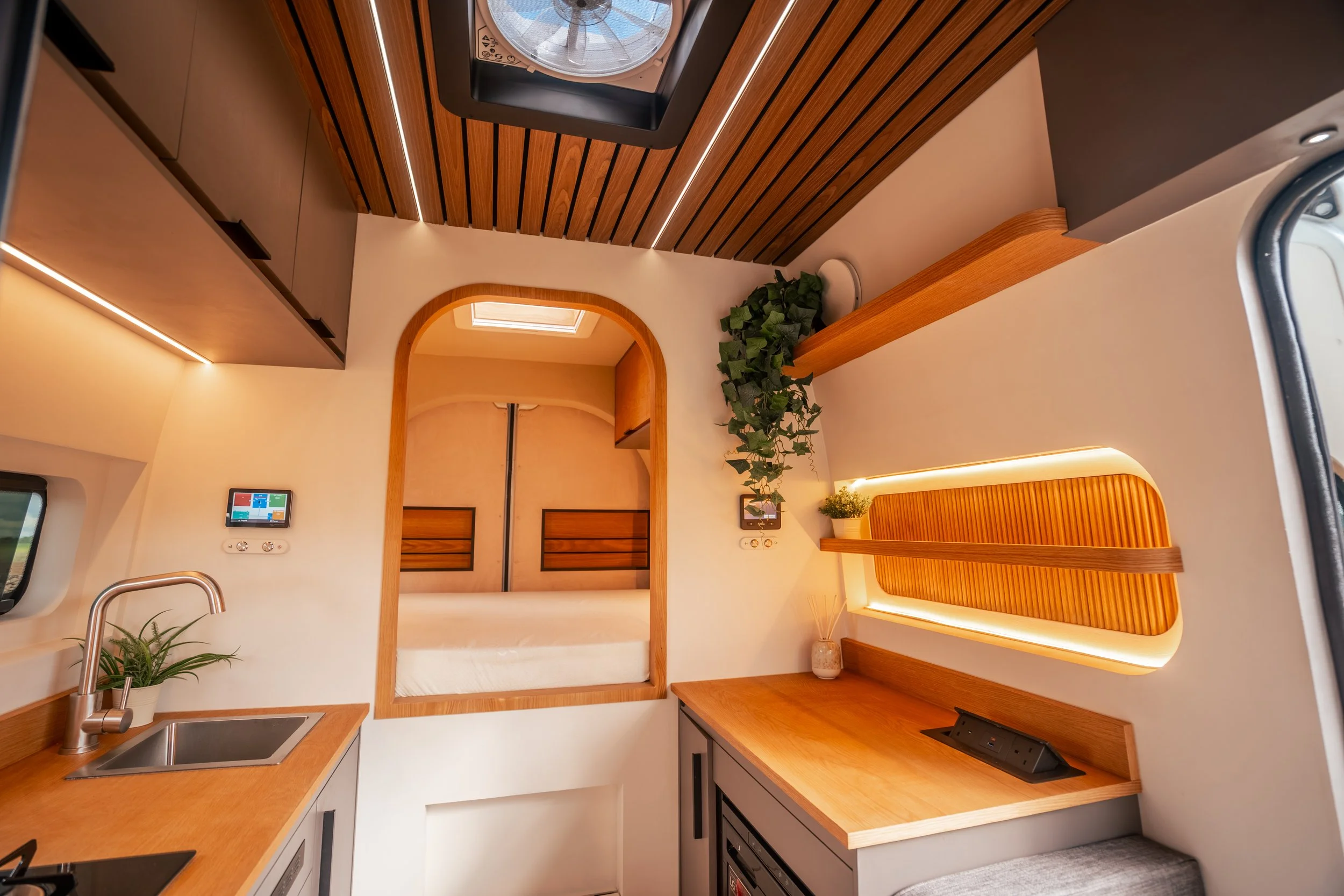 Interior of a small kitchen with wooden cabinets, a small sink with a plant in a pot, a countertop, and a wall with a skylight showing the sky. The ceiling has wooden slats with integrated lighting, and there is a small living area with a bed, a plant, and a window with sliding doors.