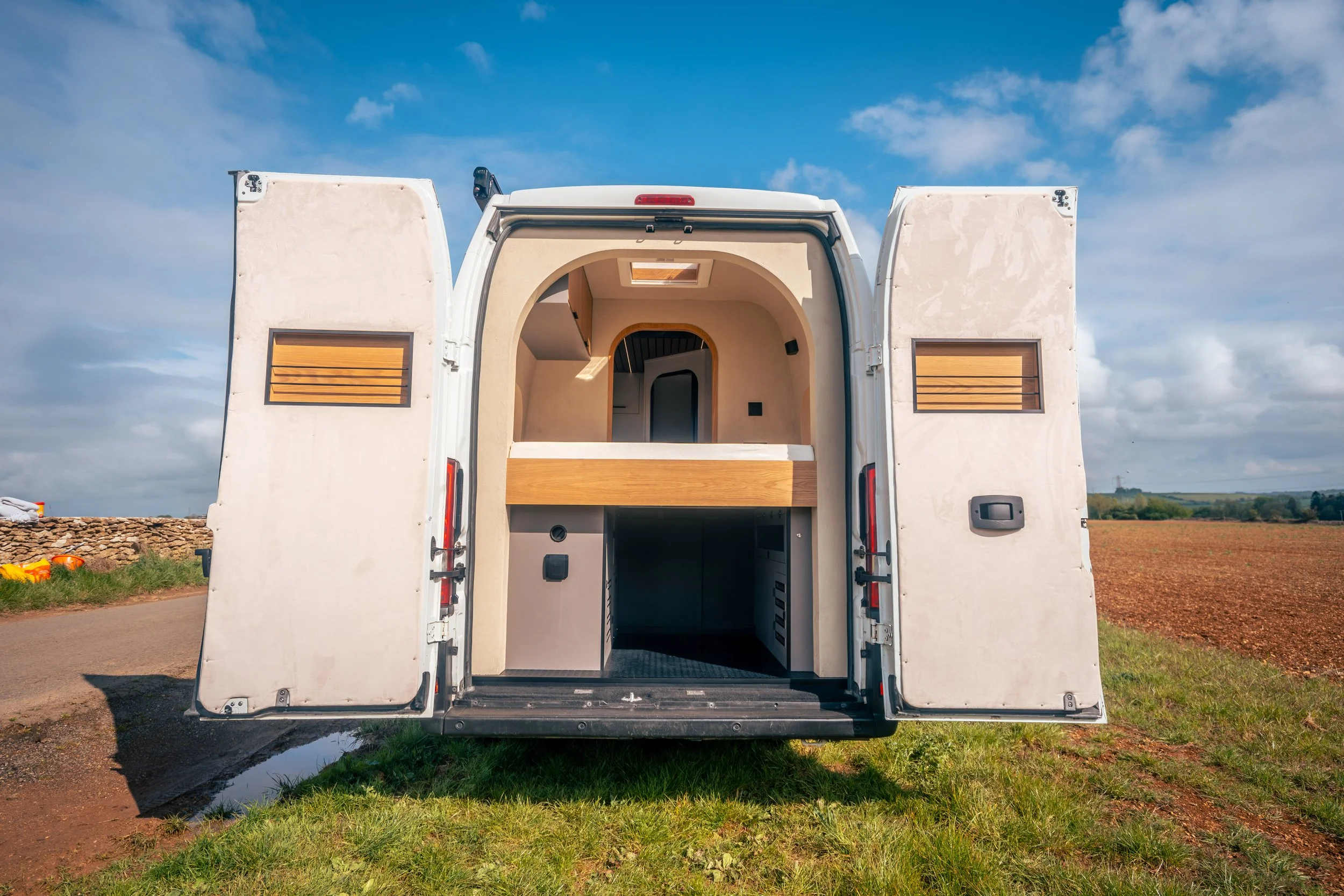 Back of a white camper van with open doors revealing an empty interior, parked on grass with a field and blue sky in the background.
