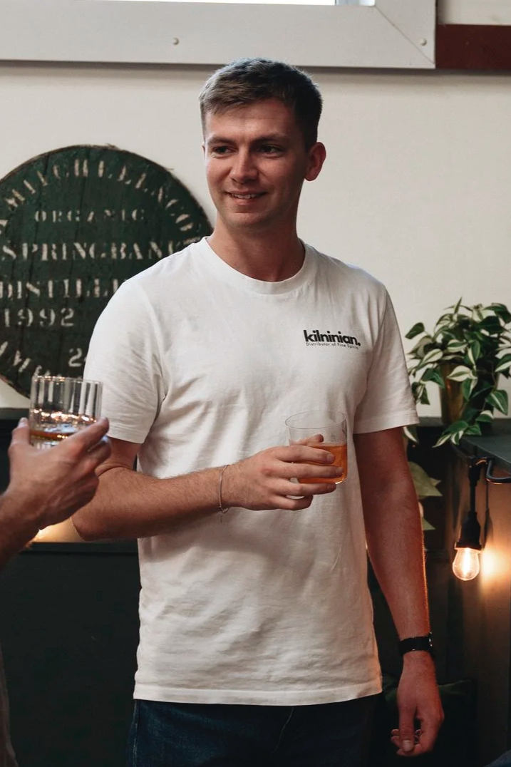 A young man with short brown hair smiling and holding a glass of amber-colored beverage at a social gathering indoors, with plants and a round decorative sign in the background.