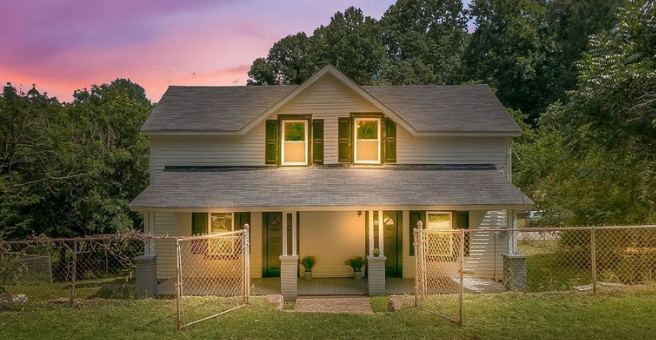 A two-story house with a porch, surrounded by a chain-link fence, and trees in the background during sunset.