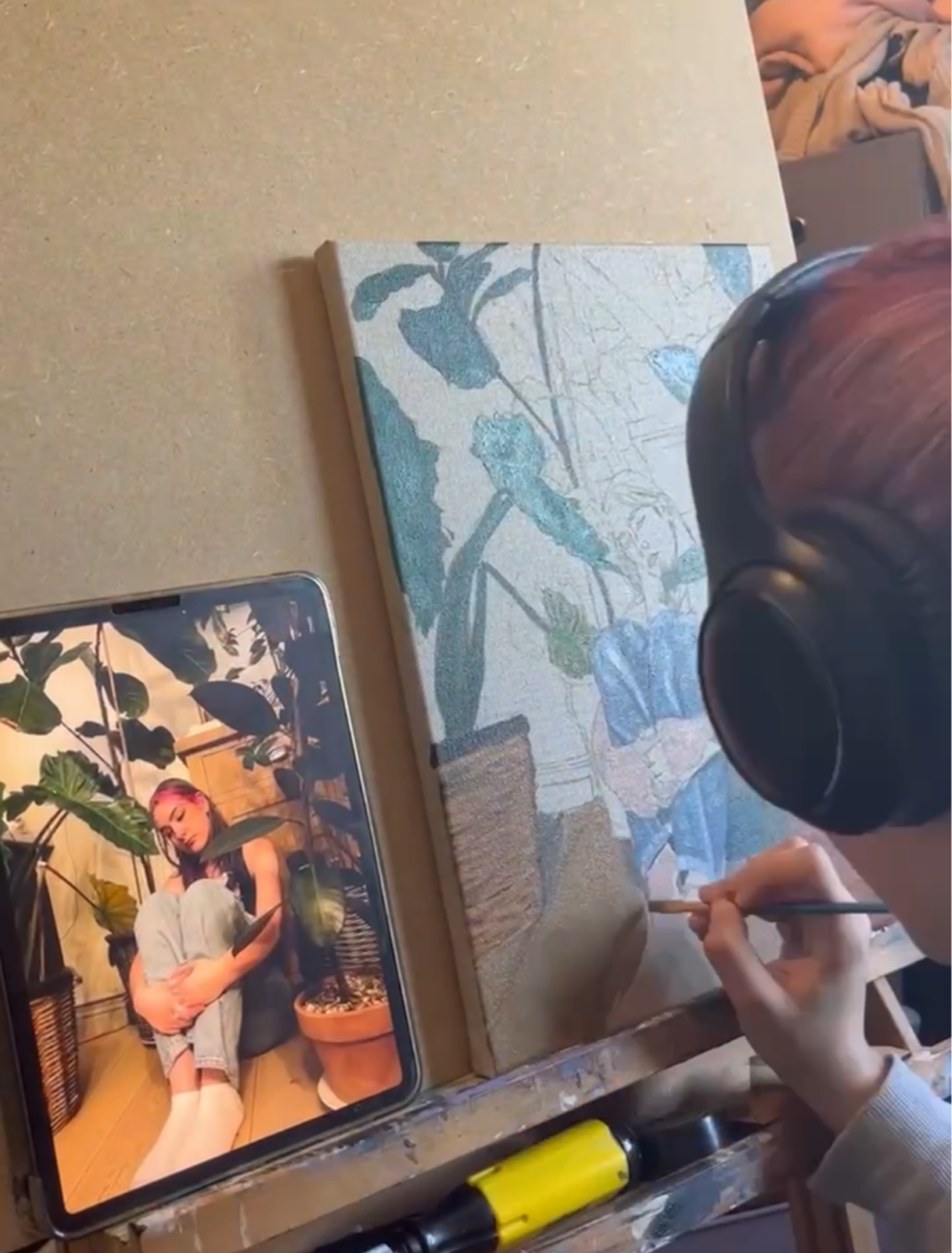 An artist is painting a detailed botanical still life on a canvas, with a photo of a woman sitting among potted plants displayed on a tablet beside the canvas.