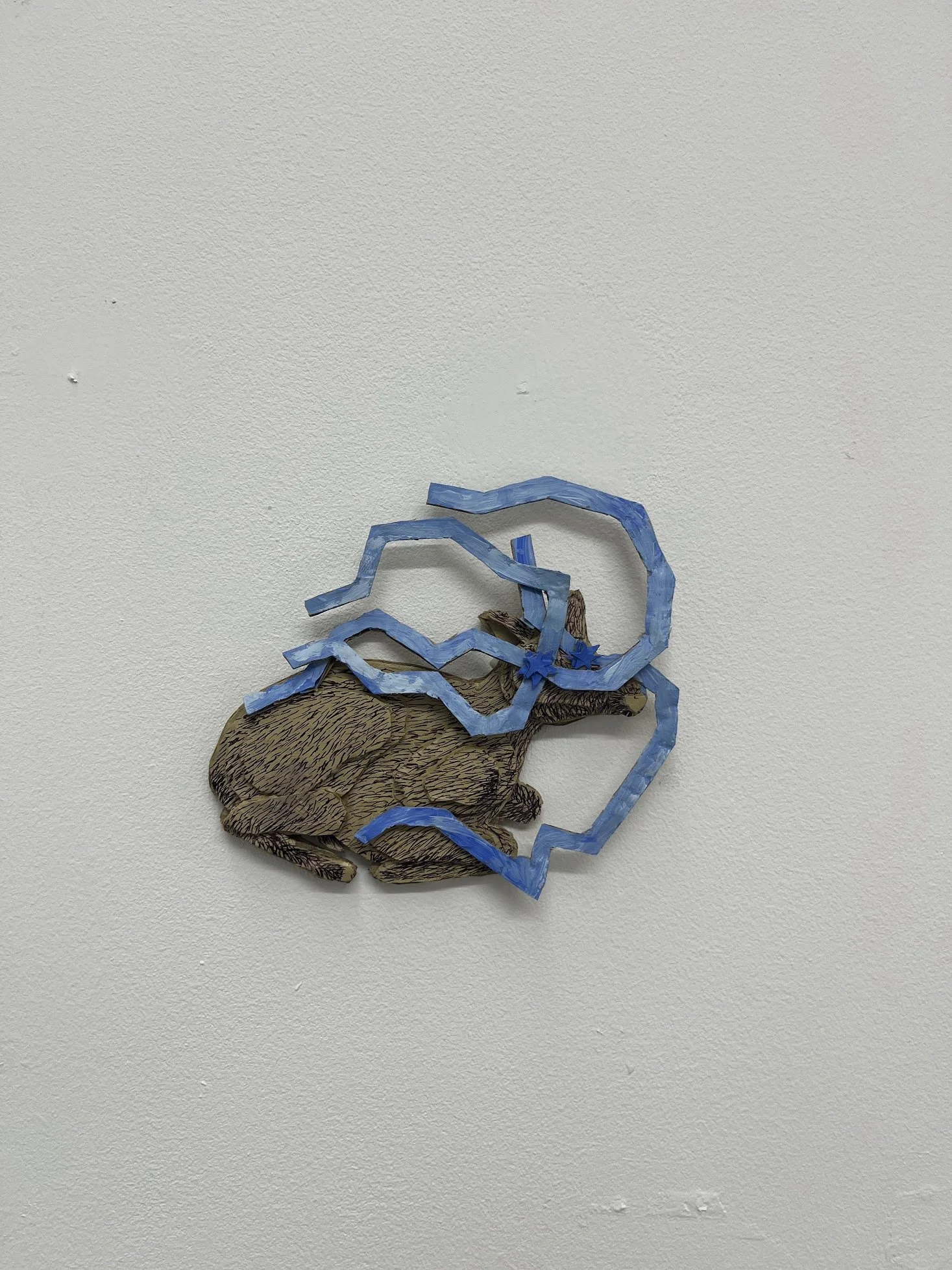 Mixed media art piece depicting a hand with a blue paper ribbon or lightning bolt shape above it, mounted on a white wall.