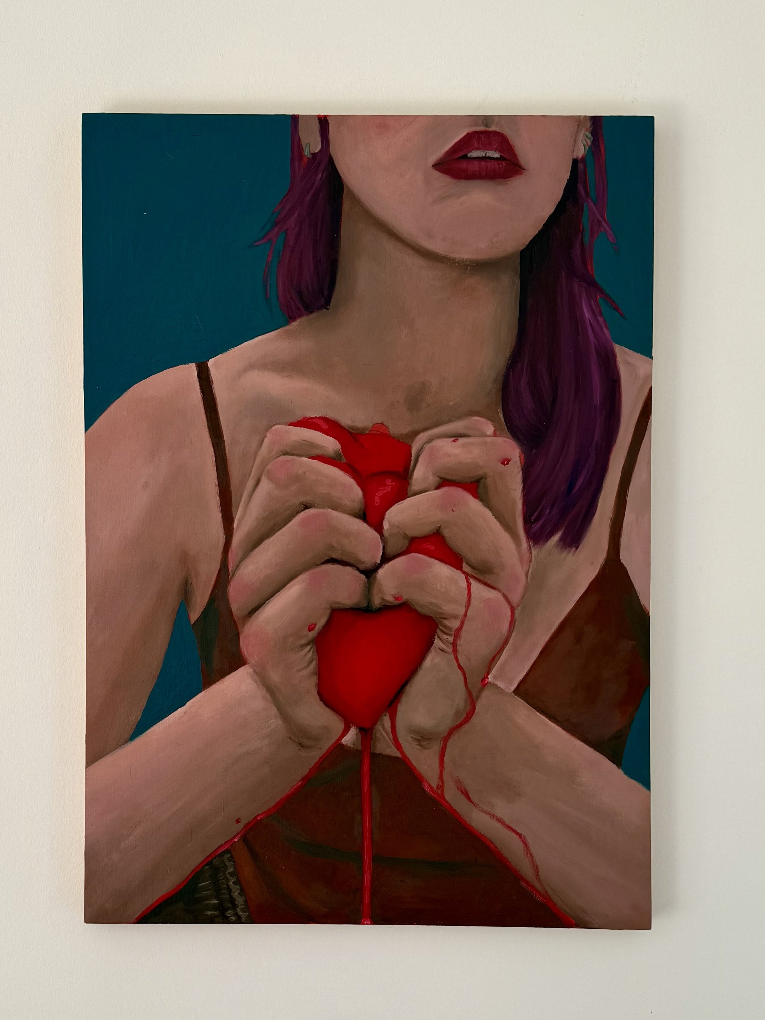 Close-up of a woman with purple hair, wearing red lipstick, holding a red heart-shaped object with red liquid. The woman is wearing a dark strap top. The background is blue.