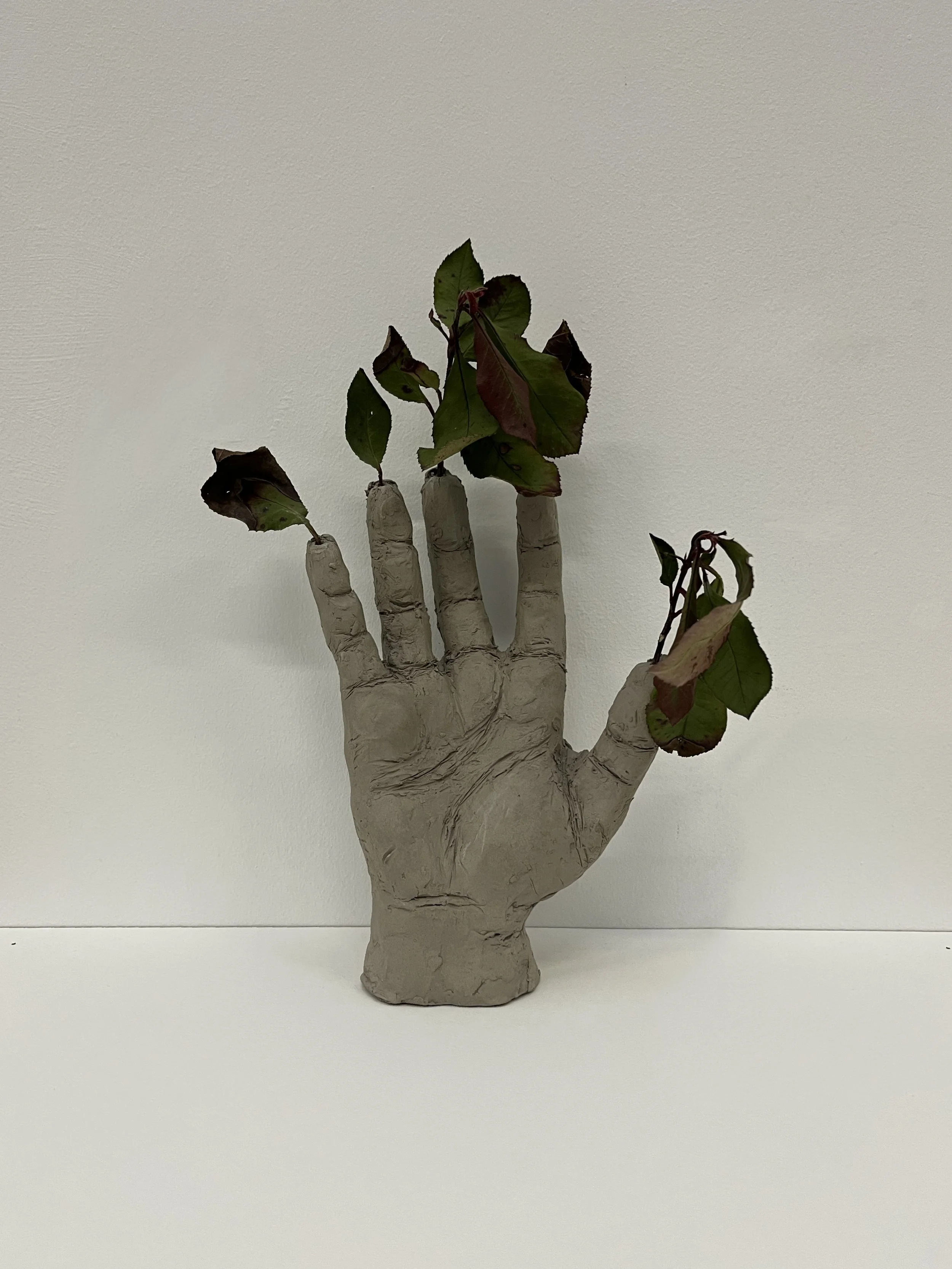 A sculpture of a human hand made to resemble tree bark, holding a small branch with green and red leaves against a plain white background.