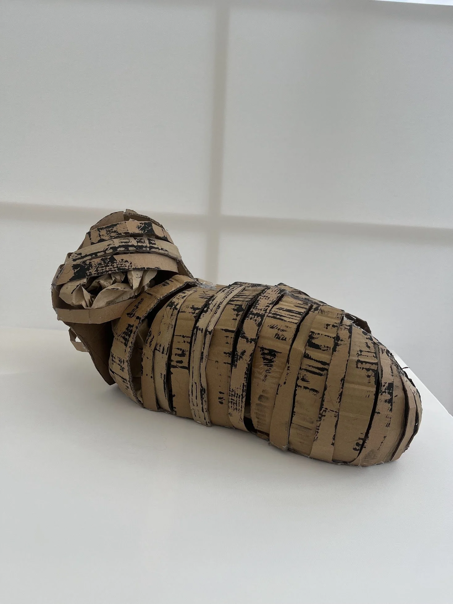 A sculpture of a curled-up snake made from crumpled and cut pieces of old cardboard with black markings, resting on a white surface against a white wall.