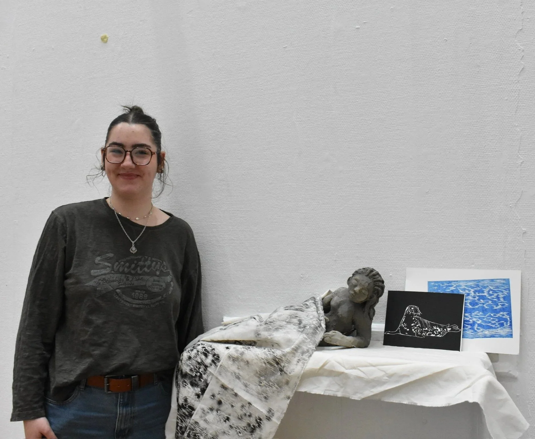 A young woman with dark hair pulled back, wearing glasses, a green T-shirt with a graphic, and a necklace, standing next to a table displaying sculptures and artwork, including a bust and a lino print of a seal.