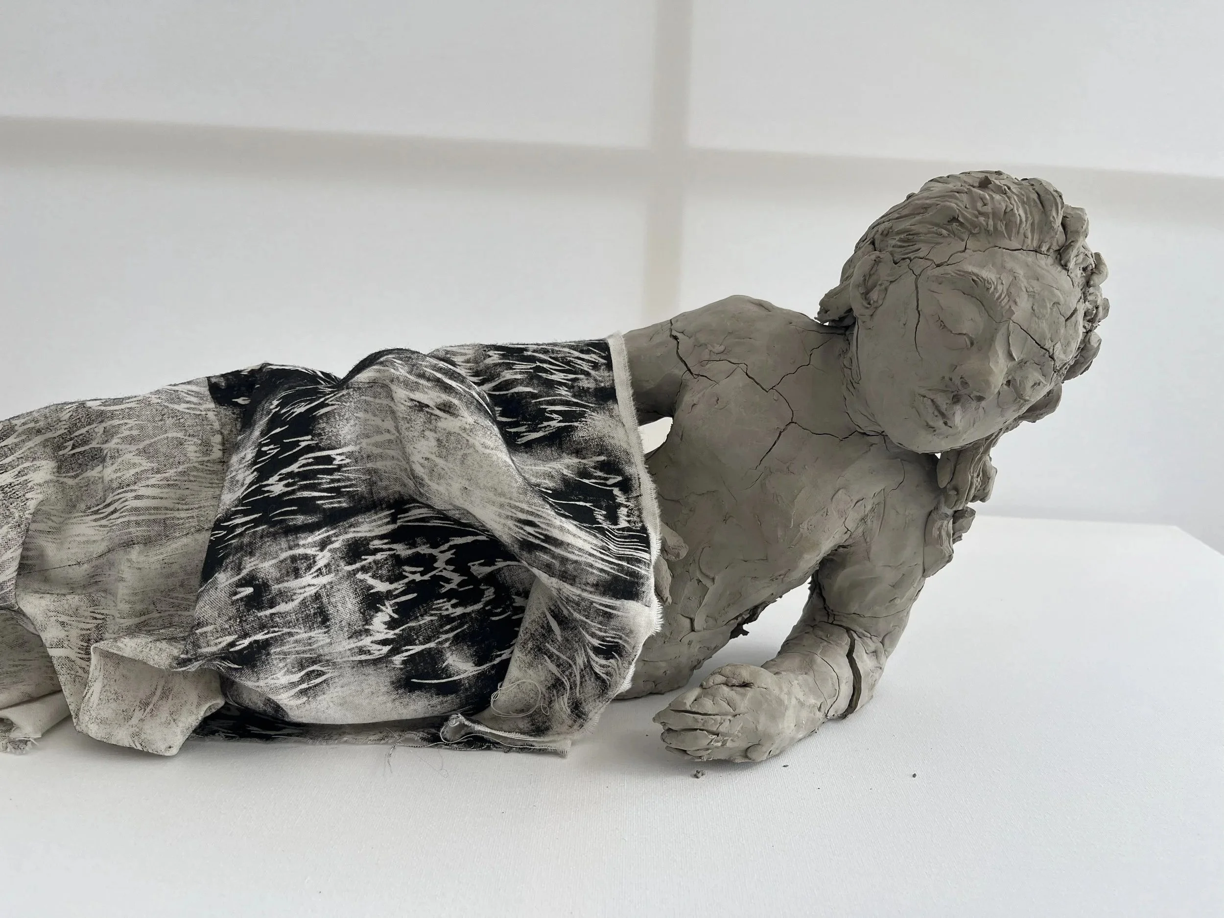 A sculpture of a seated figure with a detailed, textured face and body, covered with a cloth, placed on a white surface against a plain white background.