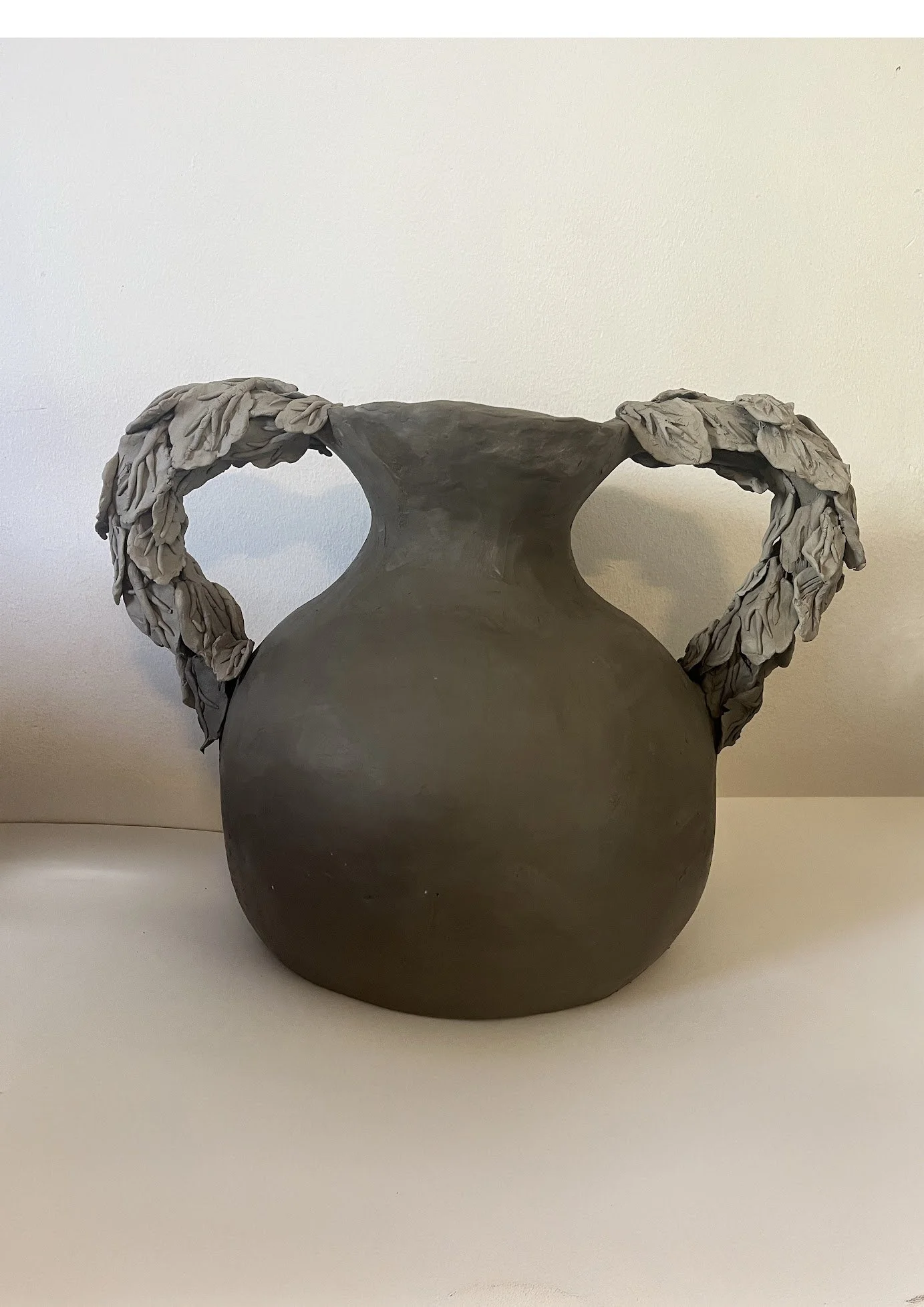 A dark gray ceramic vase with folded clay handles resembling leaves, set against a plain off-white background.