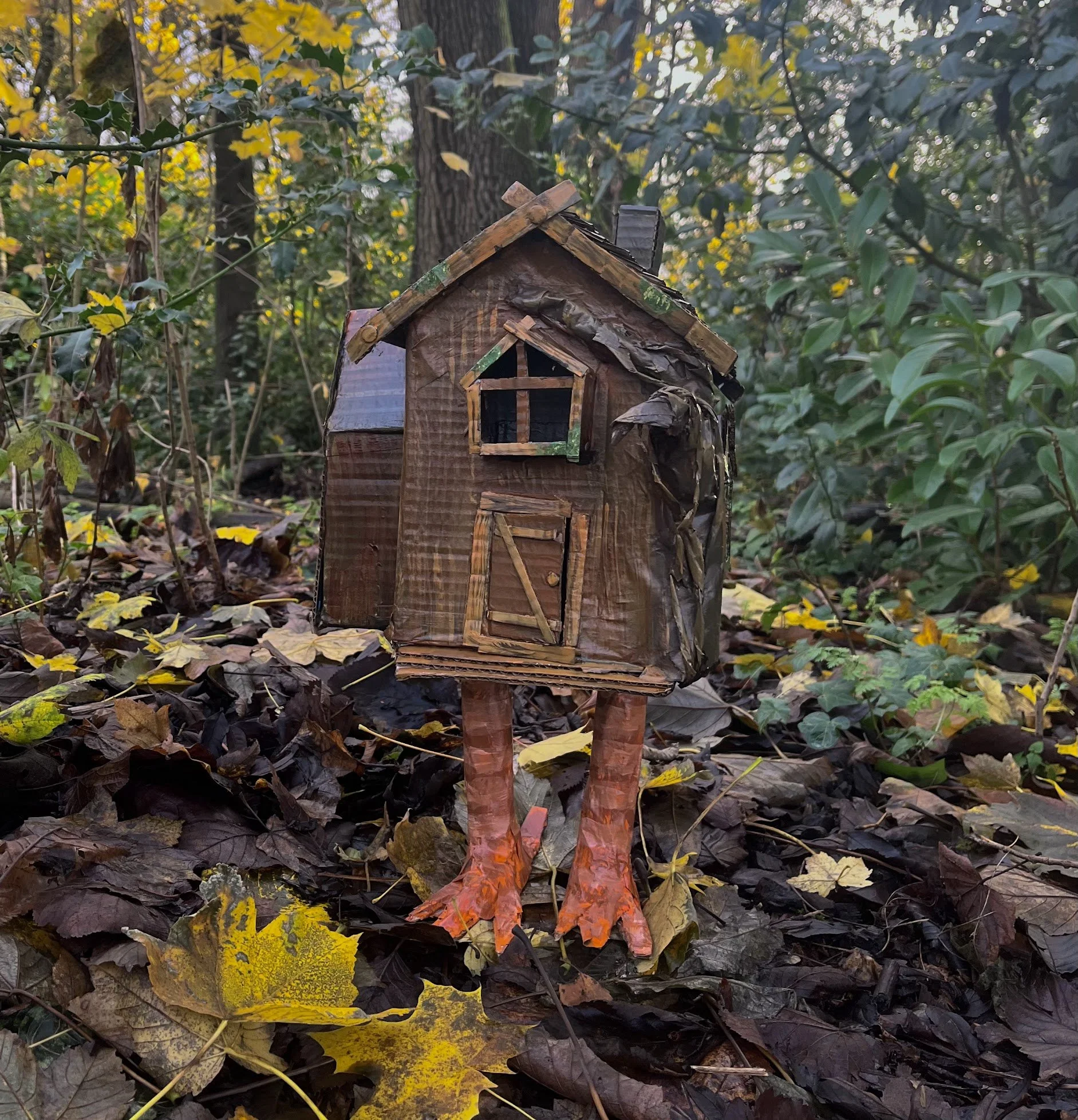 A small handmade wooden birdhouse with a triangular roof and a tiny framed window, mounted on two wooden legs among fallen autumn leaves and green shrubbery in a wooded area.
