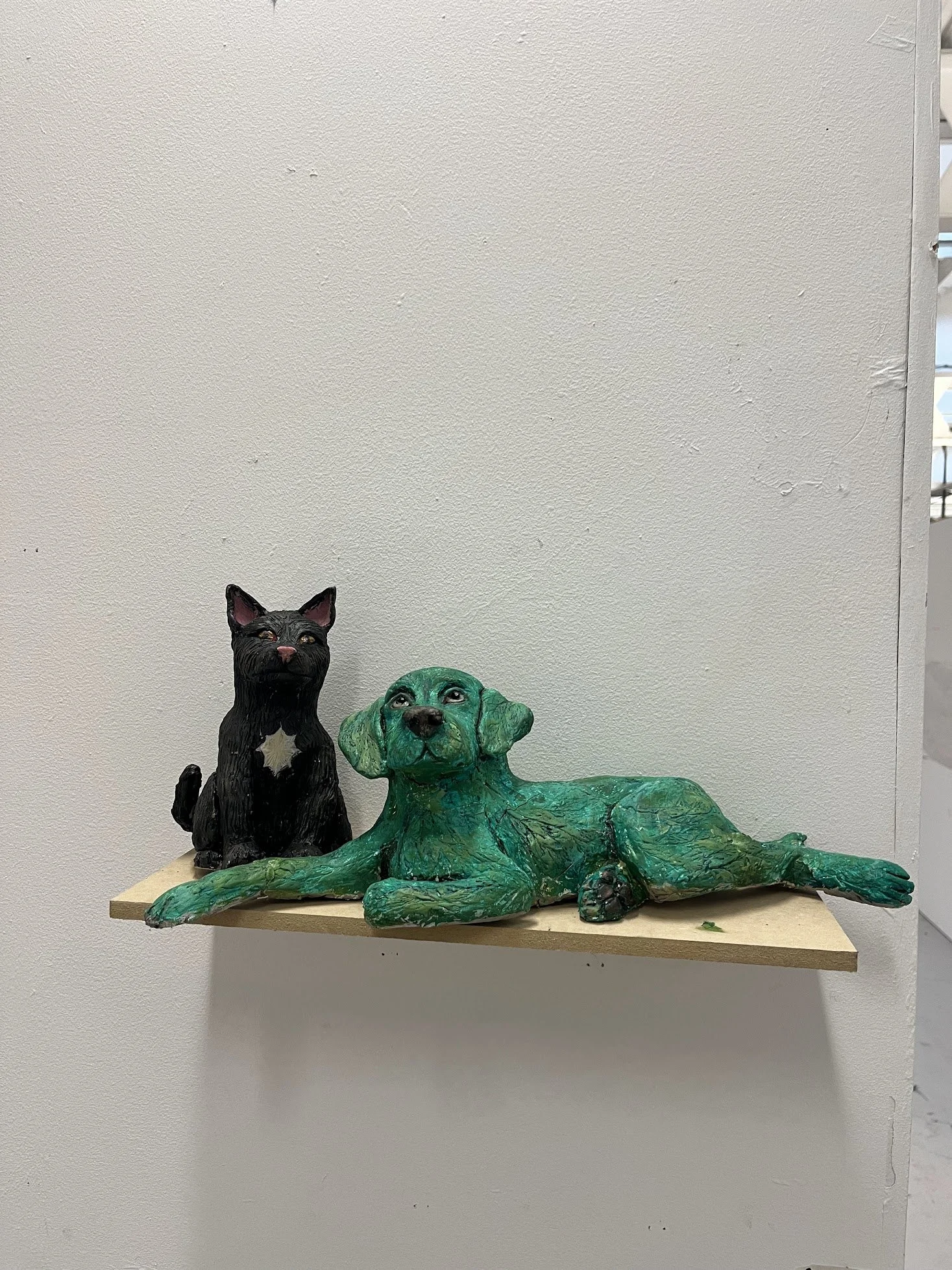 Three dog sculptures on a wooden shelf against a plain wall. One is a black dog with pointed ears sitting upright, and the other two are green dogs lying down, one with long ears and the other with floppy ears.