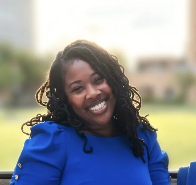A woman with long, curly black hair and a big smile, wearing a bright blue top, outdoors with a blurred background of greenery and sunlight.