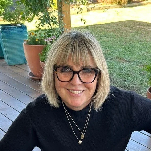 A woman with blonde hair and glasses smiling outdoors on a wooden deck, with potted plants and a grassy yard in the background.