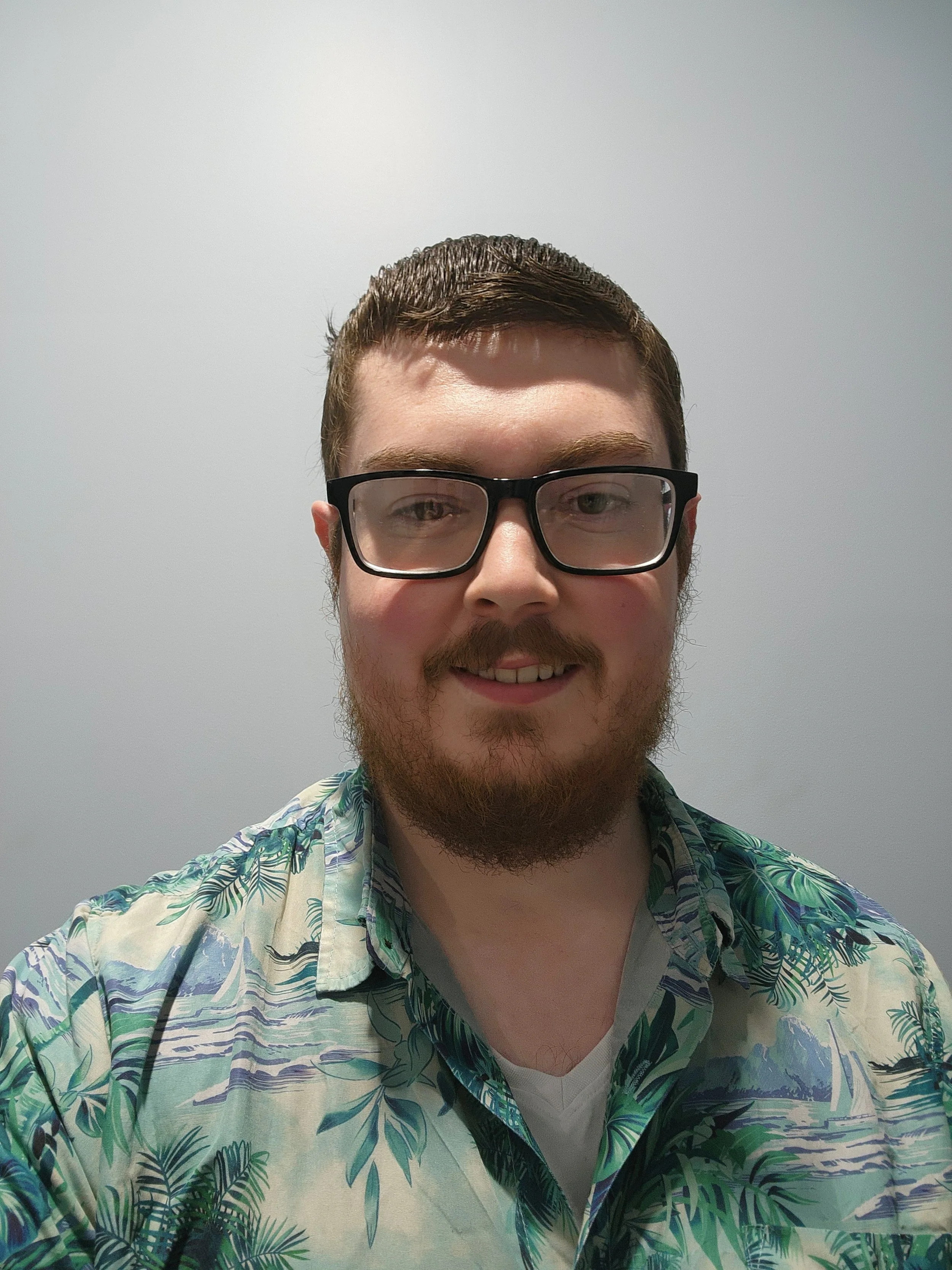 A young man with short brown hair, glasses, a beard, and a mustache smiling in front of a plain, light-colored wall. He is wearing a tropical-patterned shirt.