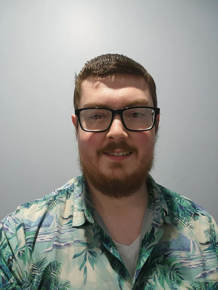 A young man with short brown hair, glasses, a beard and a moustache, smiling in front of a plain, light-coloured wall. He is wearing a tropical-patterned shirt.