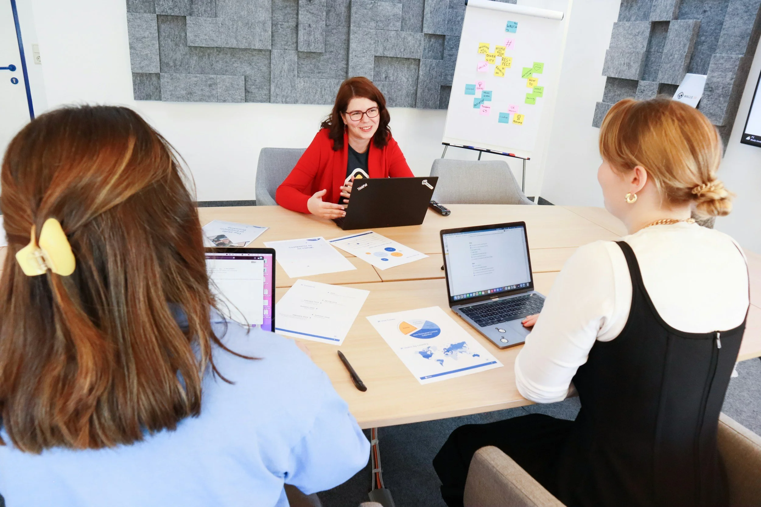 Four women seated around a conference table working on laptops, with papers and charts, in a meeting room with gray acoustic panels and a whiteboard with sticky notes.