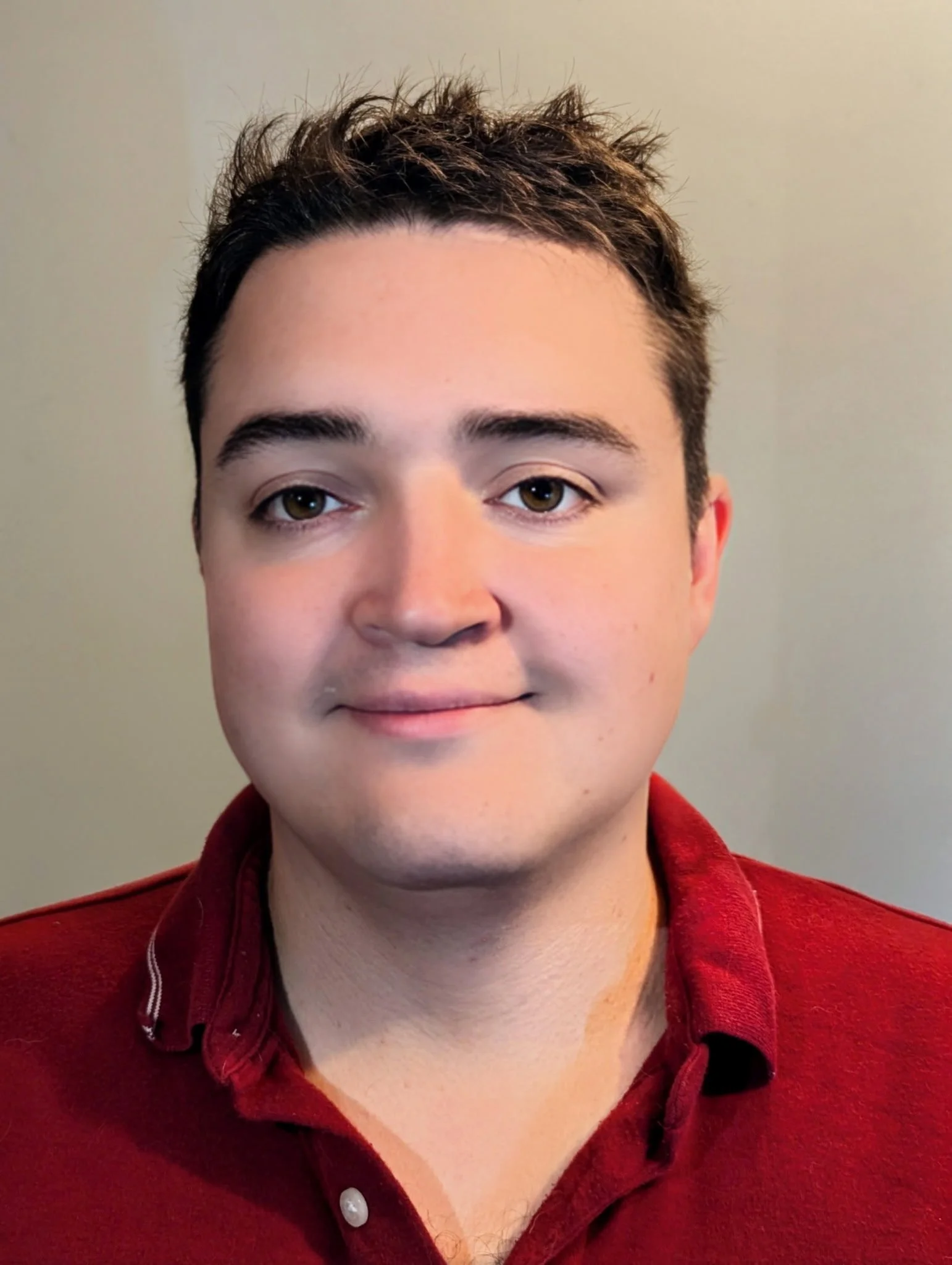 Close-up photo of a young man with short, dark curly hair, brown eyes, light skin, wearing a red shirt with a collar, against a plain beige background.
