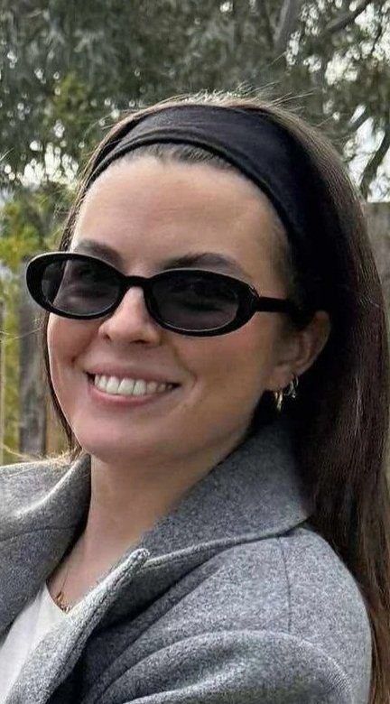 A woman with long dark hair, wearing black sunglasses, a black headband, earrings, and a gray blazer, smiling outdoors with trees in the background.